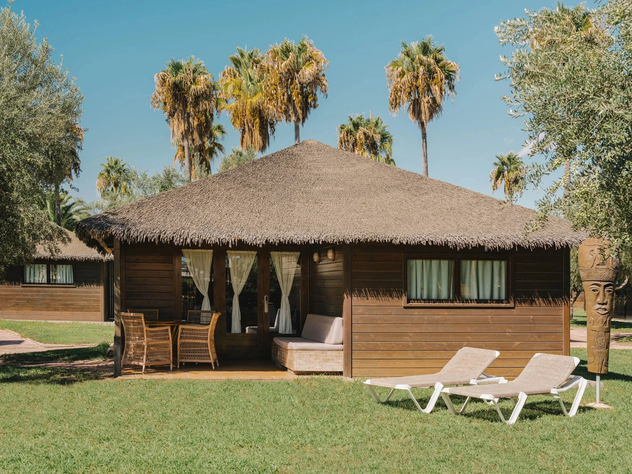 A thatched-roof bungalow with dark wooden exterior, surrounded by palm trees and greenery, featuring outdoor seating and lounge chairs.