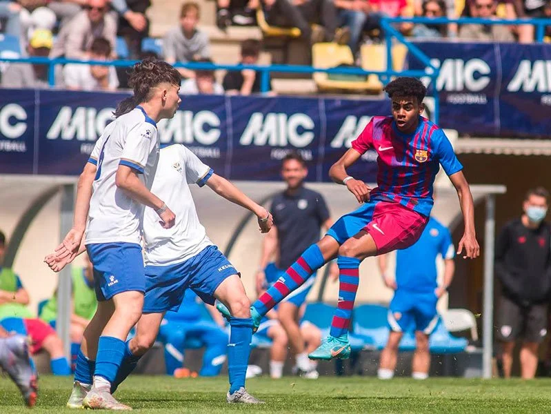 Soccer match with players from FC Barcelona and another team, with one player in mid-air kicking the ball during a game with spectators in the background.
