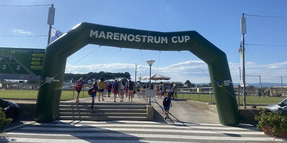Group of children and adults entering a sports field through an inflatable archway that reads 'Marenostrum Cup' under a blue sky.