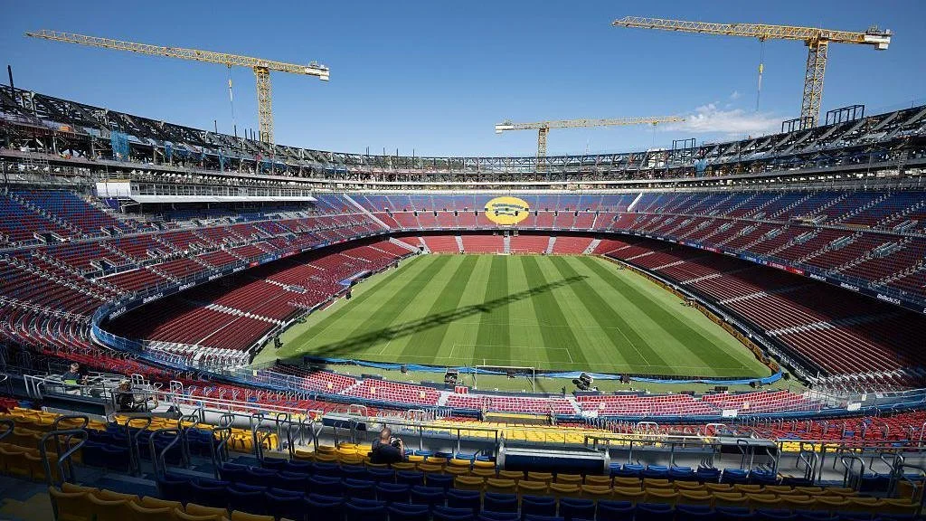 Empty soccer stadium with green field, red and blue seats, construction cranes overhead, and clear blue sky.