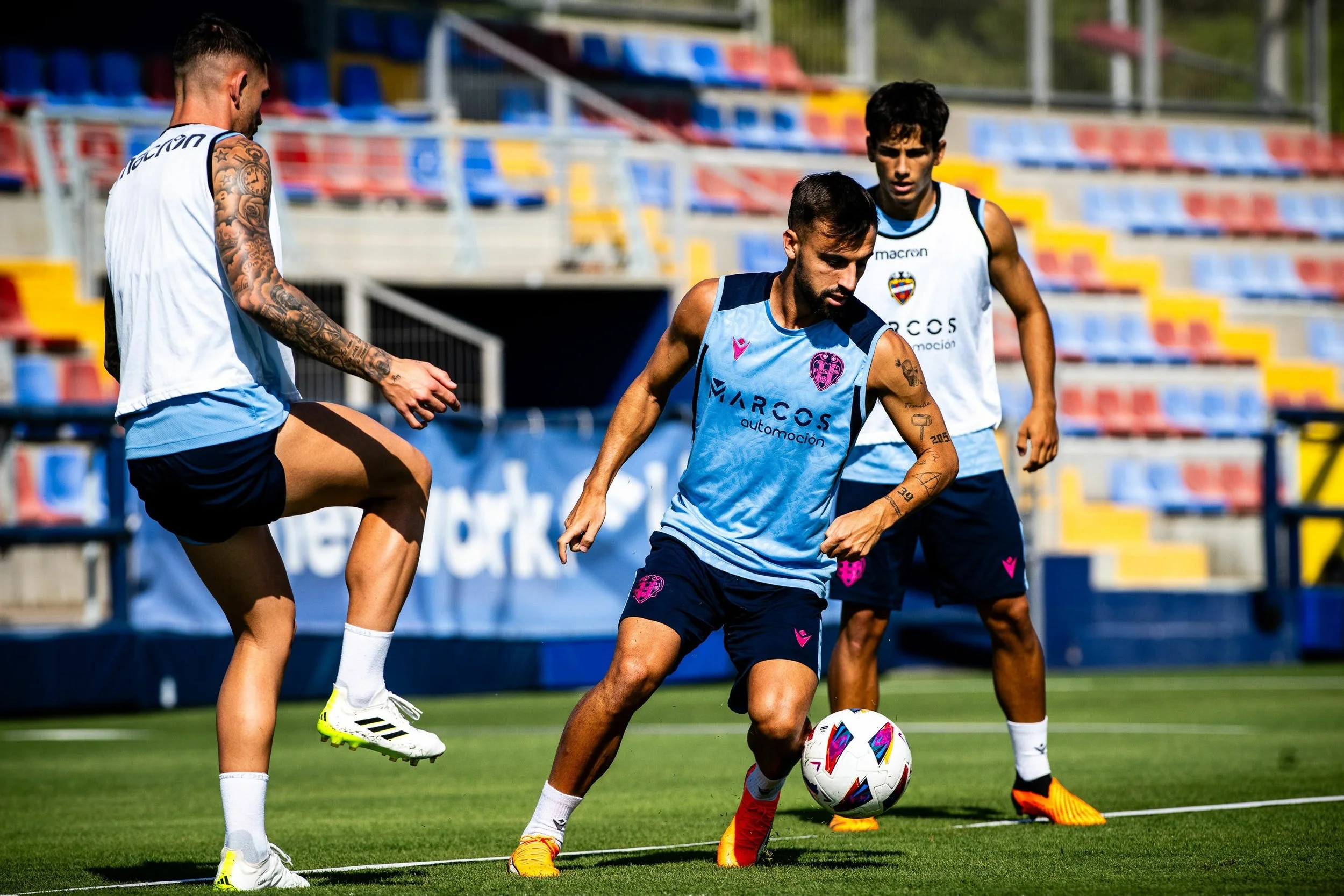 Three male soccer players practicing on a field, with two in white and blue uniforms and one in a light blue jersey and shorts, playing with a soccer ball during daytime.