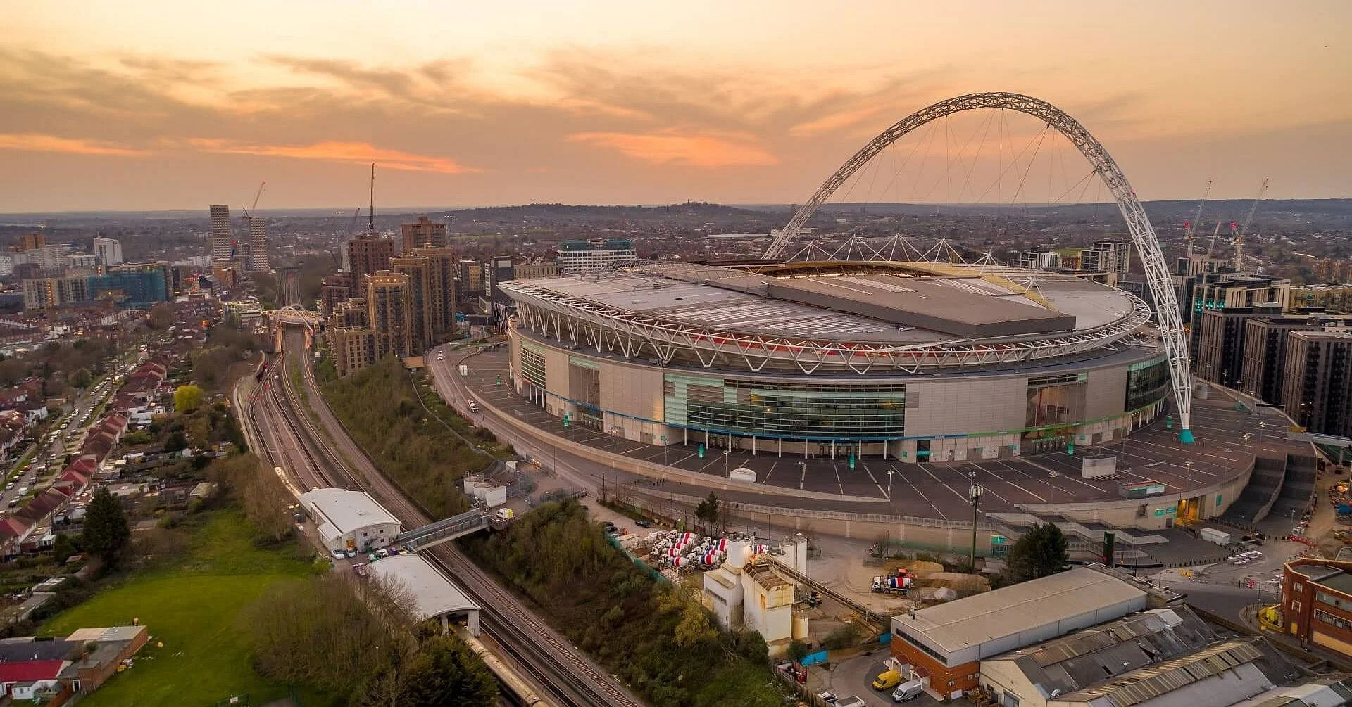 An aerial view of a large modern stadium with a distinctive arch, set in an urban area at sunset. The stadium features a partially covered roof, numerous surrounding buildings, roads, and railway tracks.