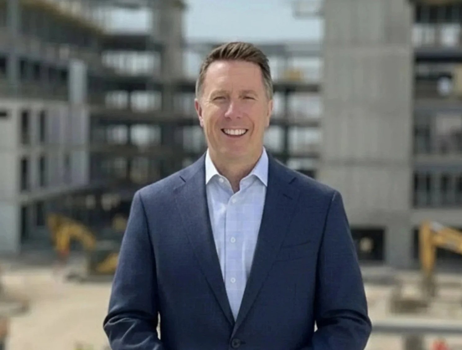 Ash Rich standing outdoors in business attire with a commercial construction project in the background.