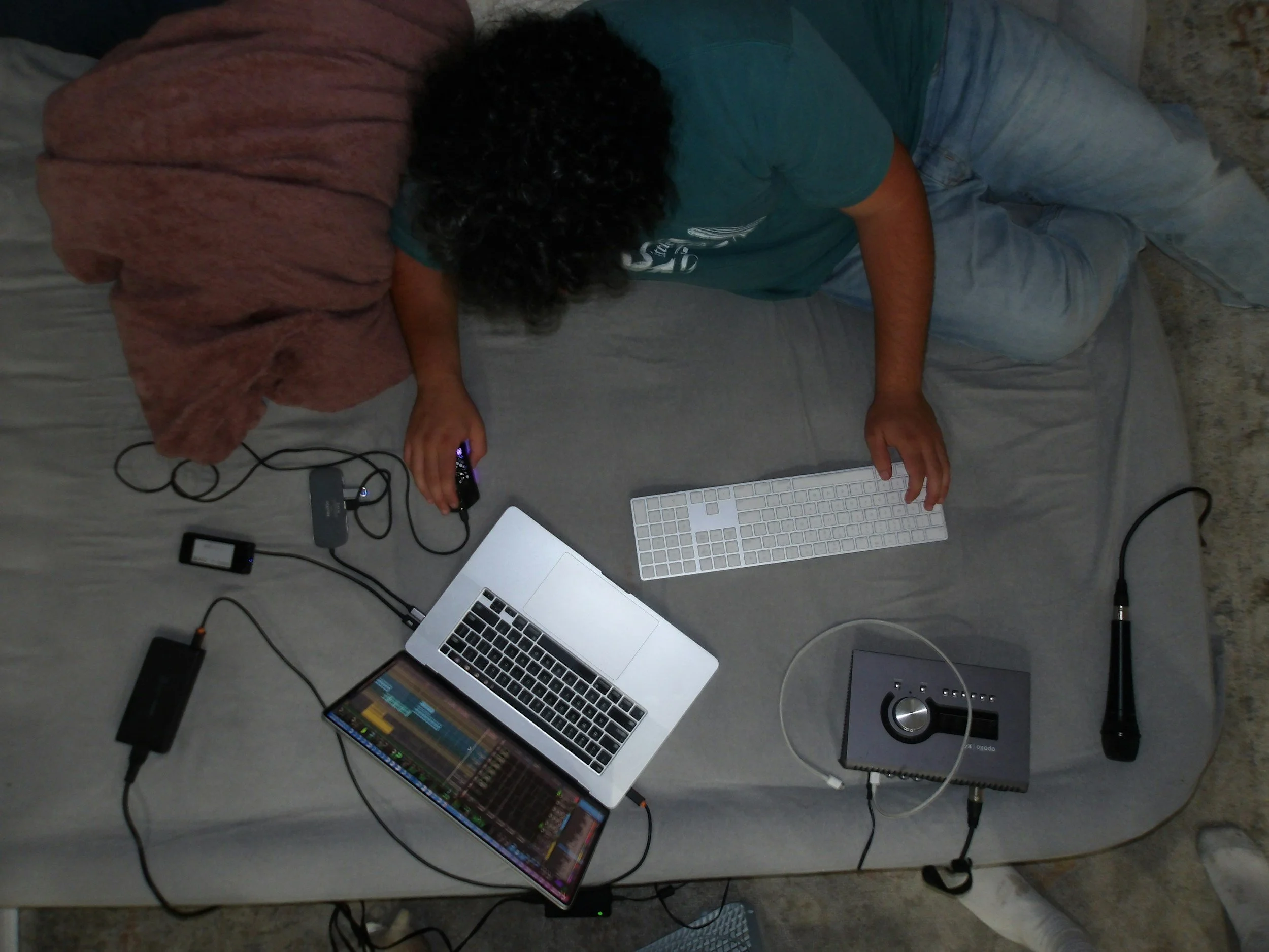 Person with curly hair lying on a bed, working with electronic music equipment including a laptop, MIDI keyboard, audio interface, and a microphone.