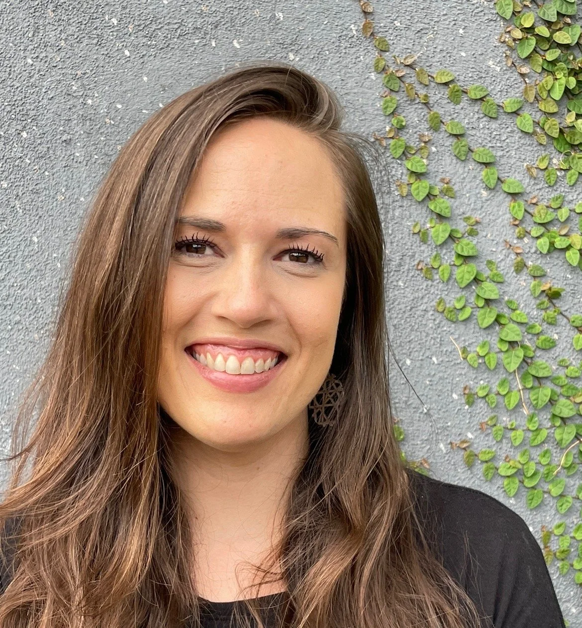 Close-up of a smiling woman with long brown hair standing against a textured gray wall with green ivy.