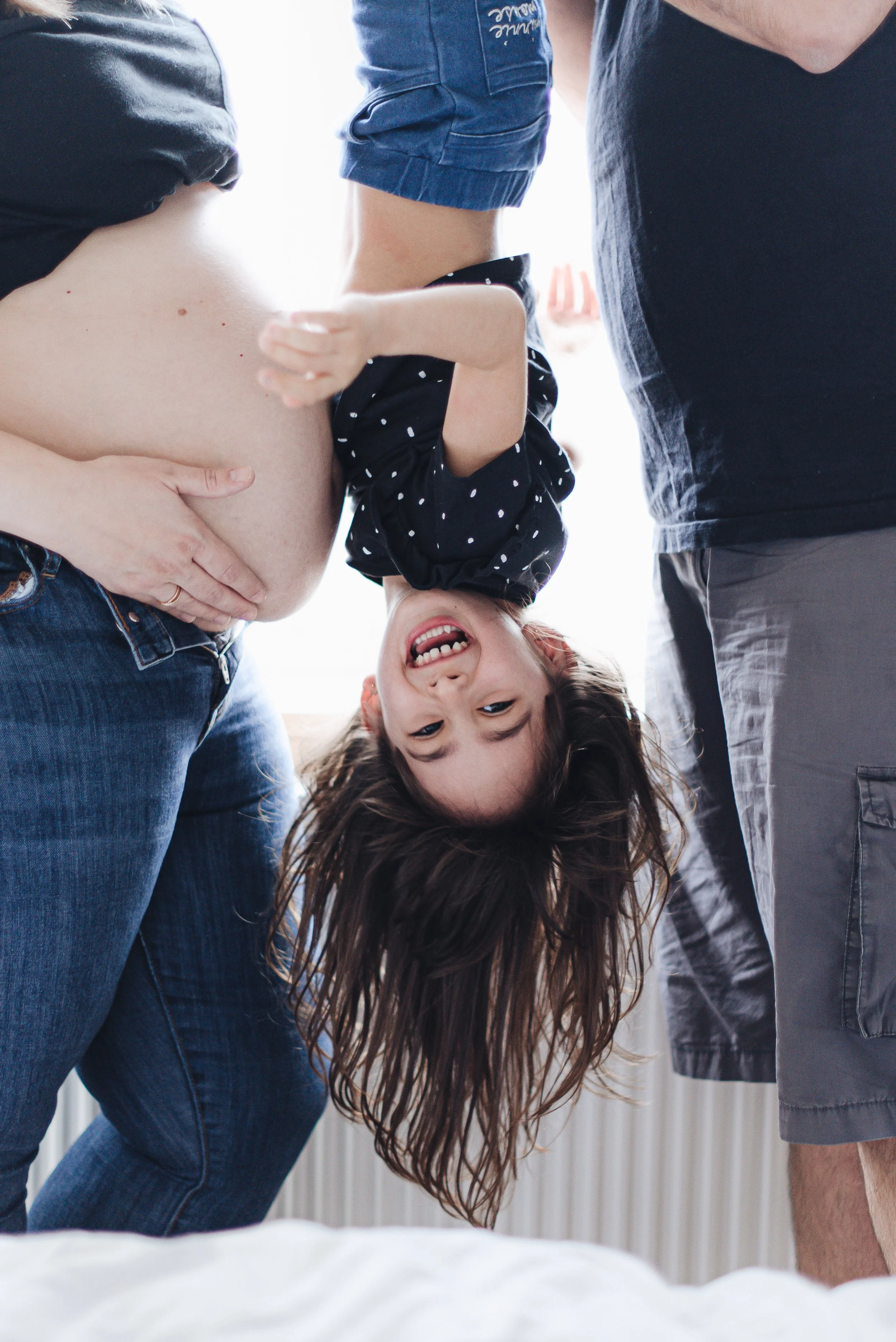 A happy young girl hanging upside down between two adults, smiling and giggling during a playful moment in a bright room.
