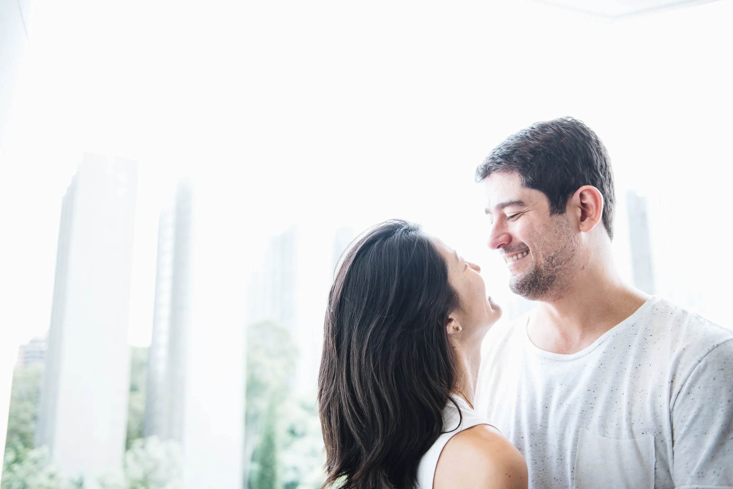 Family photoshoot with a couple smiling and looking at each other outdoors with city buildings in the background.