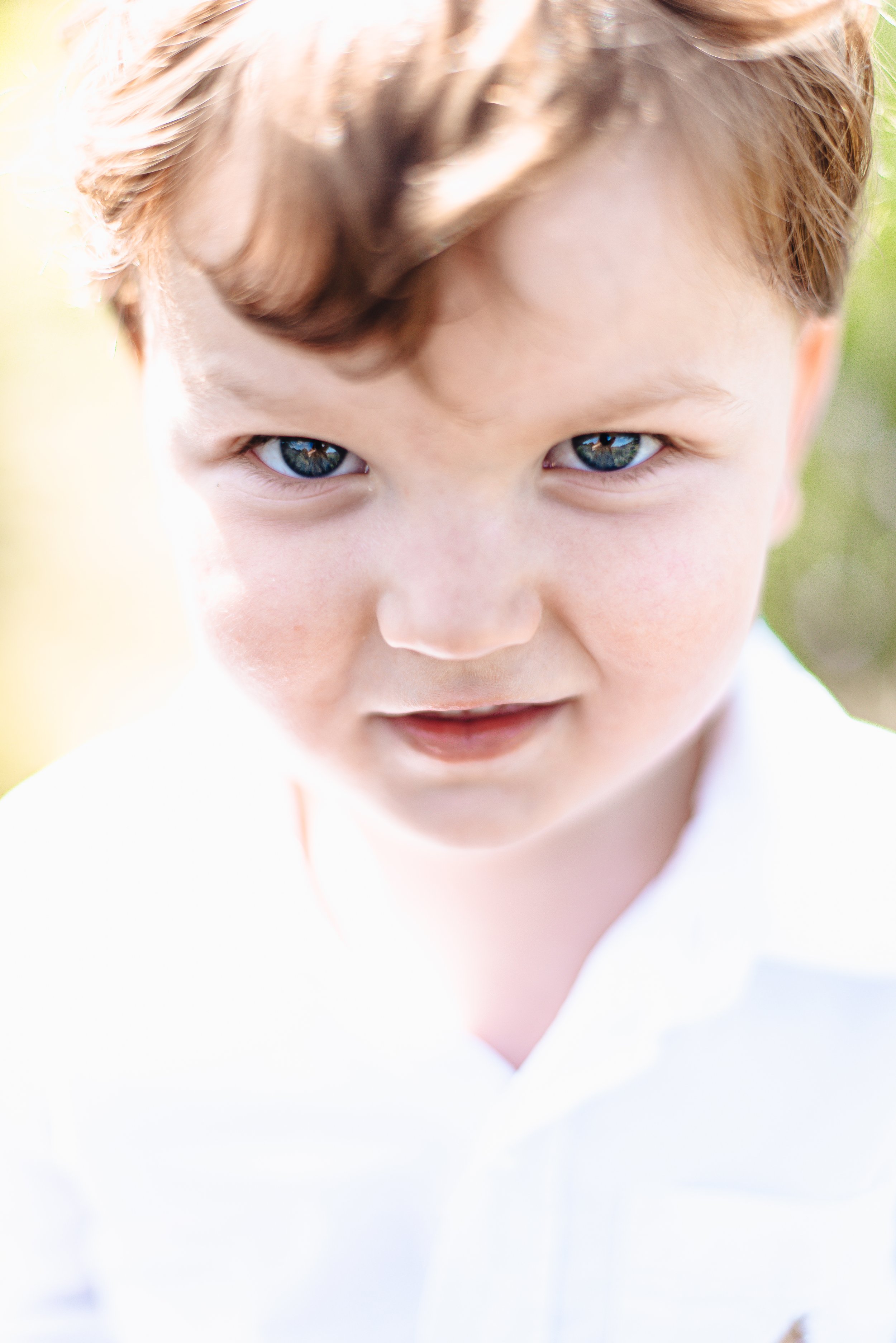 Close-up of a young boy with blue eyes and brown hair, wearing a white shirt, outdoors with sunlight.