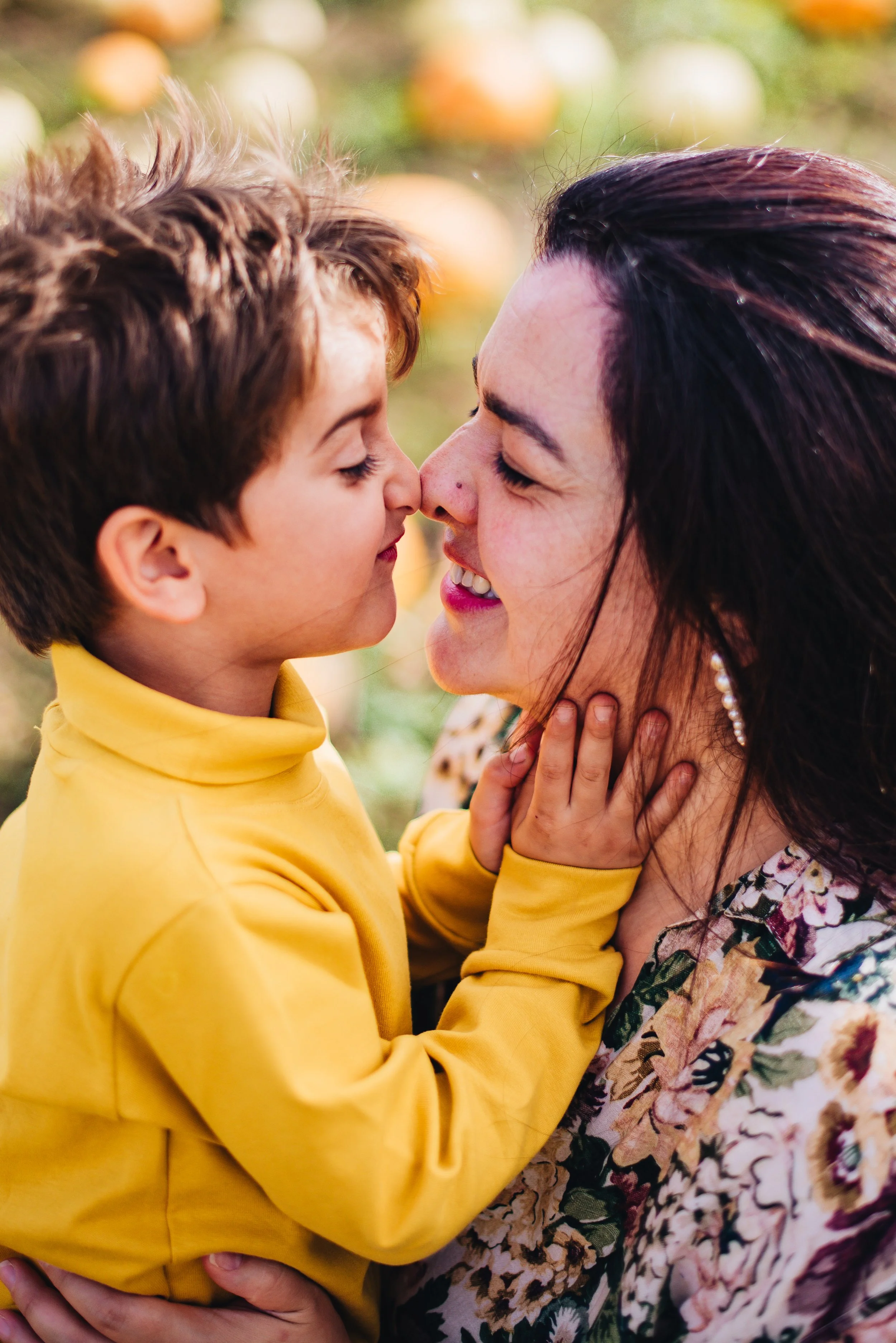 A young boy and a woman are touching noses and smiling at each other outdoors, with a blurred background of pumpkin decorations.