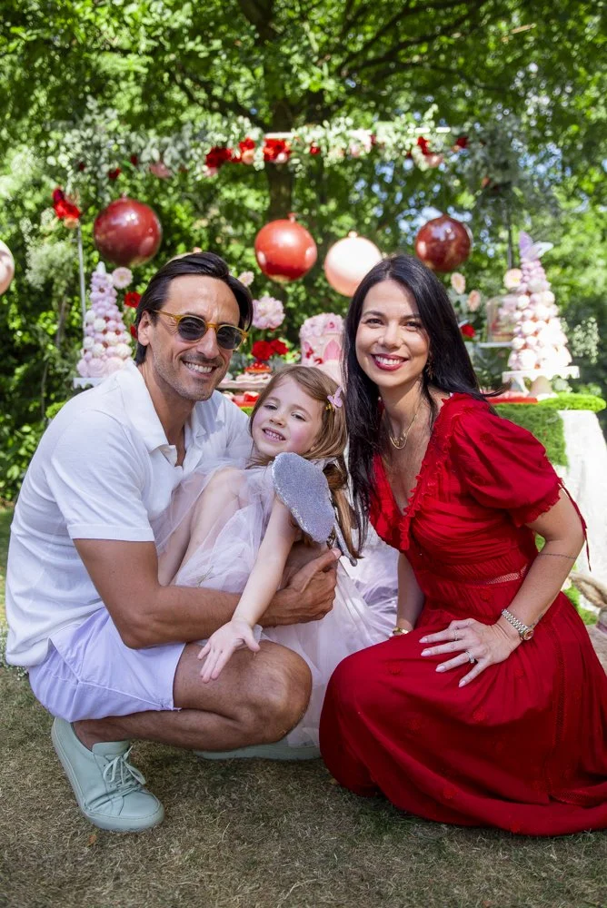 A happy family of three posing outdoors at a birthday celebration, with a decorated table and trees in the background. The man is wearing a white shirt and shorts, the woman in a red dress, and the girl in a light pink dress with glittery wings.
