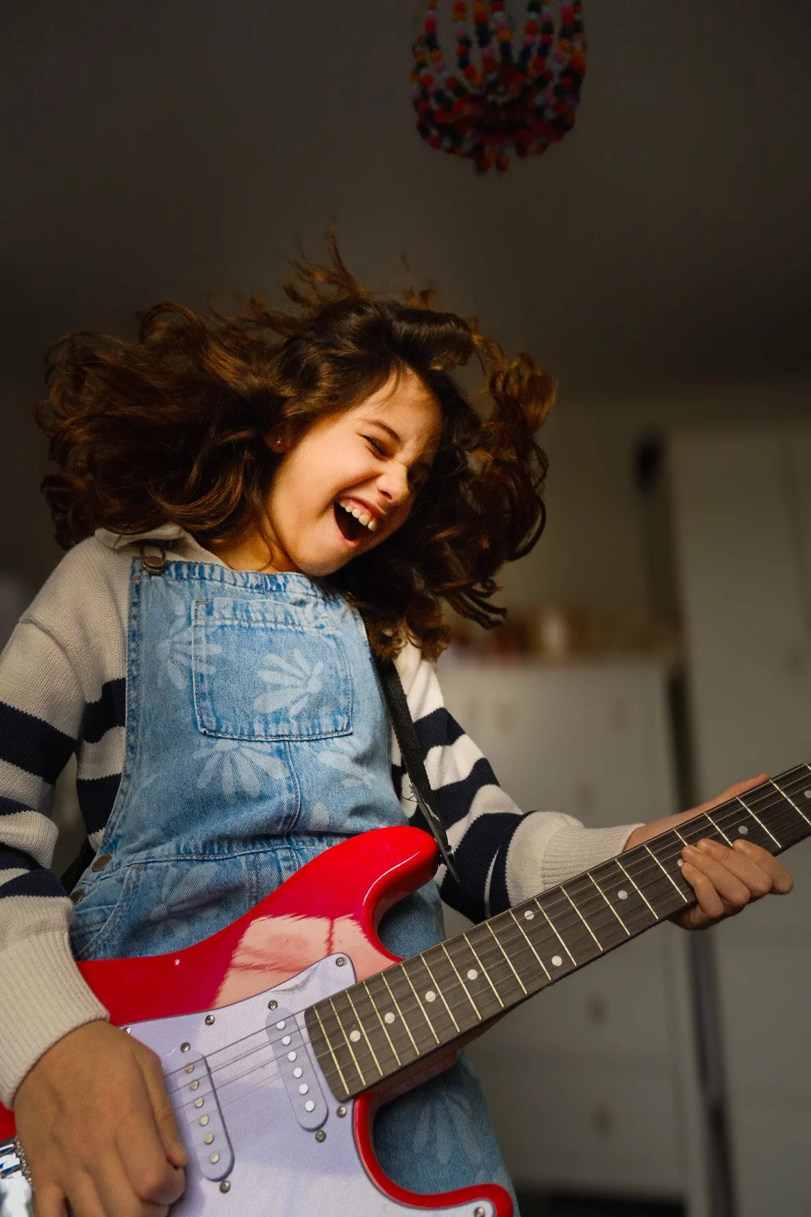 A young girl with curly brown hair wearing a striped sweater and denim overalls, smiling and playing a red electric guitar.