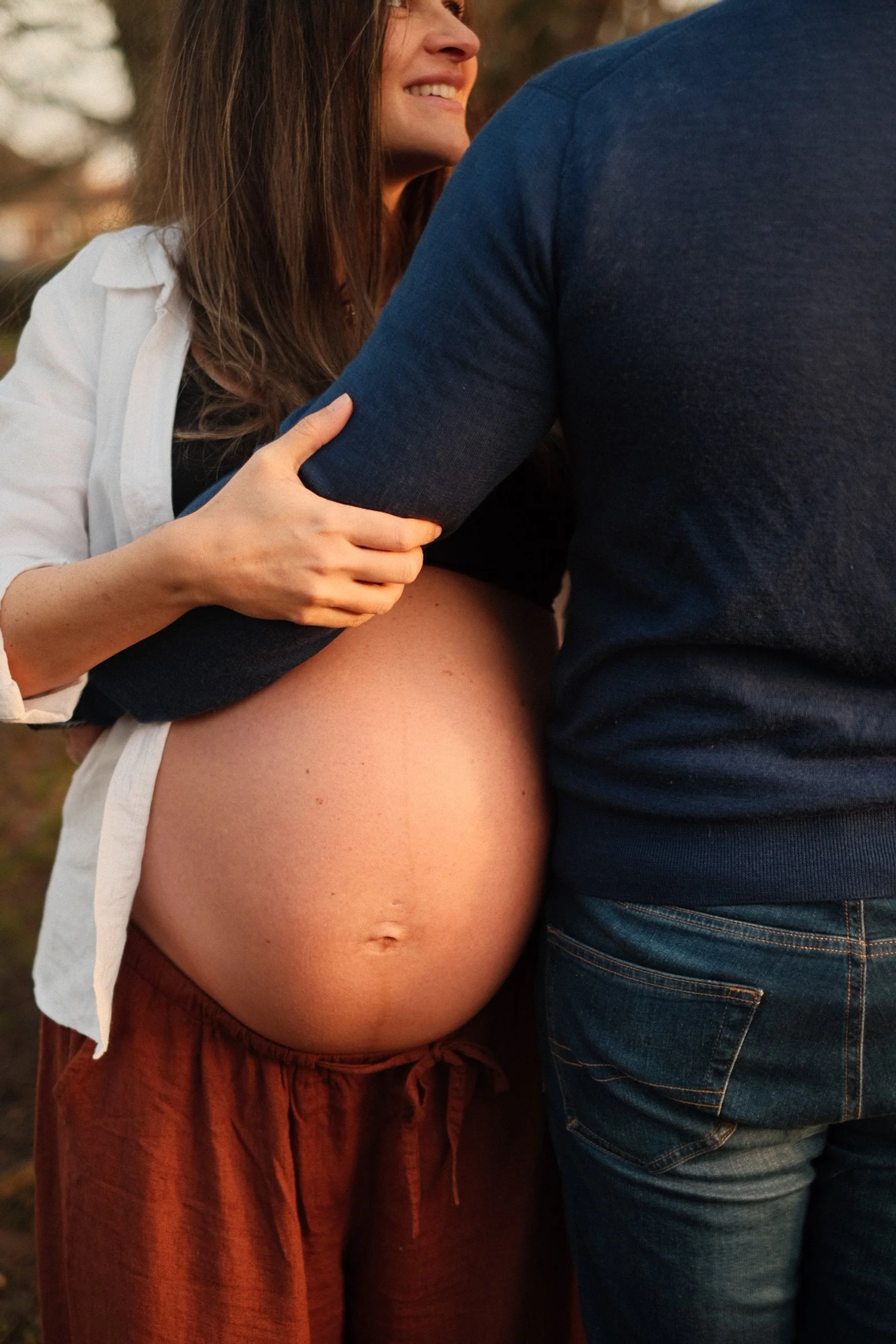 A pregnant woman smiling, with her hand on a man's arm, in an outdoor setting during sunset.