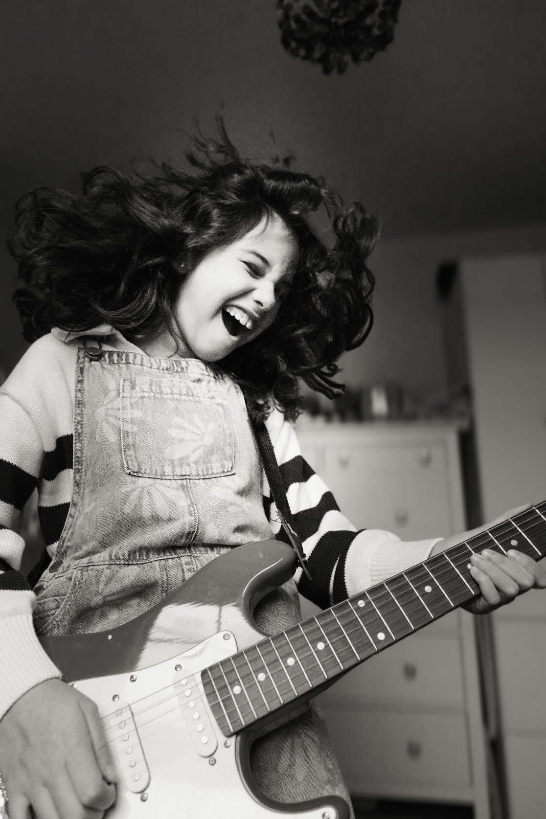 A young woman with curly hair playing an electric guitar, smiling and laughing, in a joyful moment indoors.
