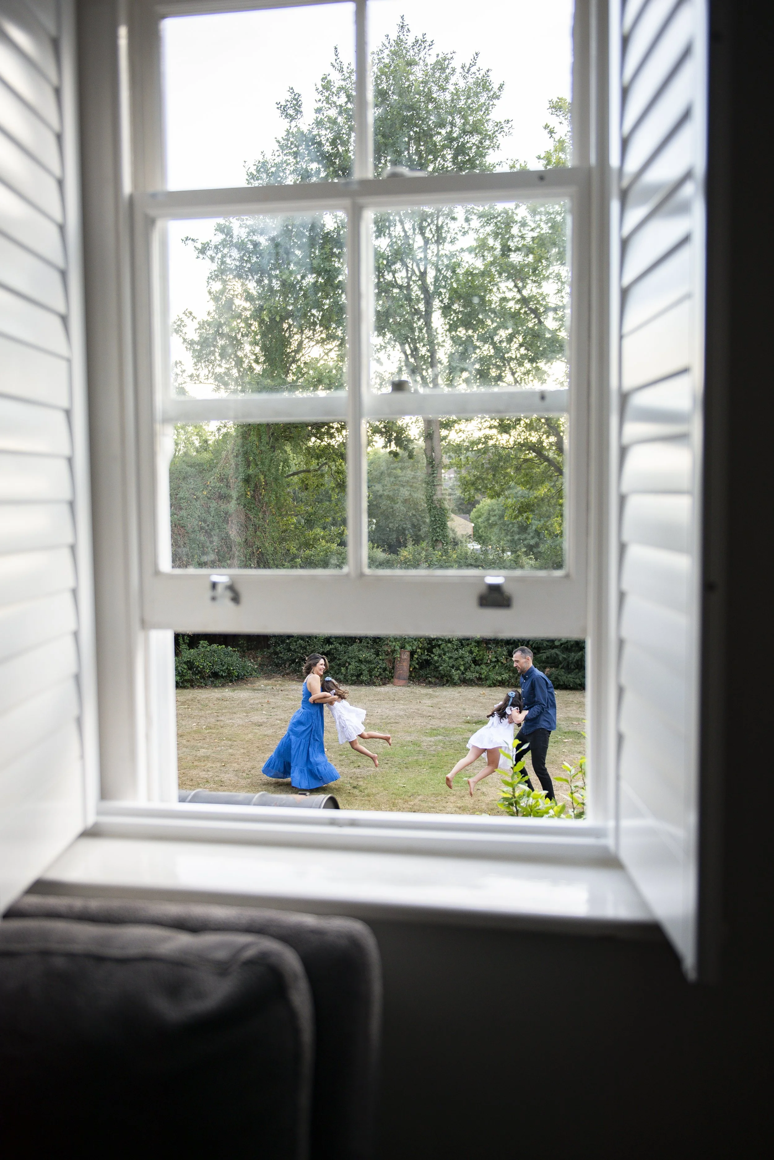 View of a family outside through a window with open shutters, a woman in a blue dress and two young girls in white dresses playing and running on a grassy lawn during daytime.
