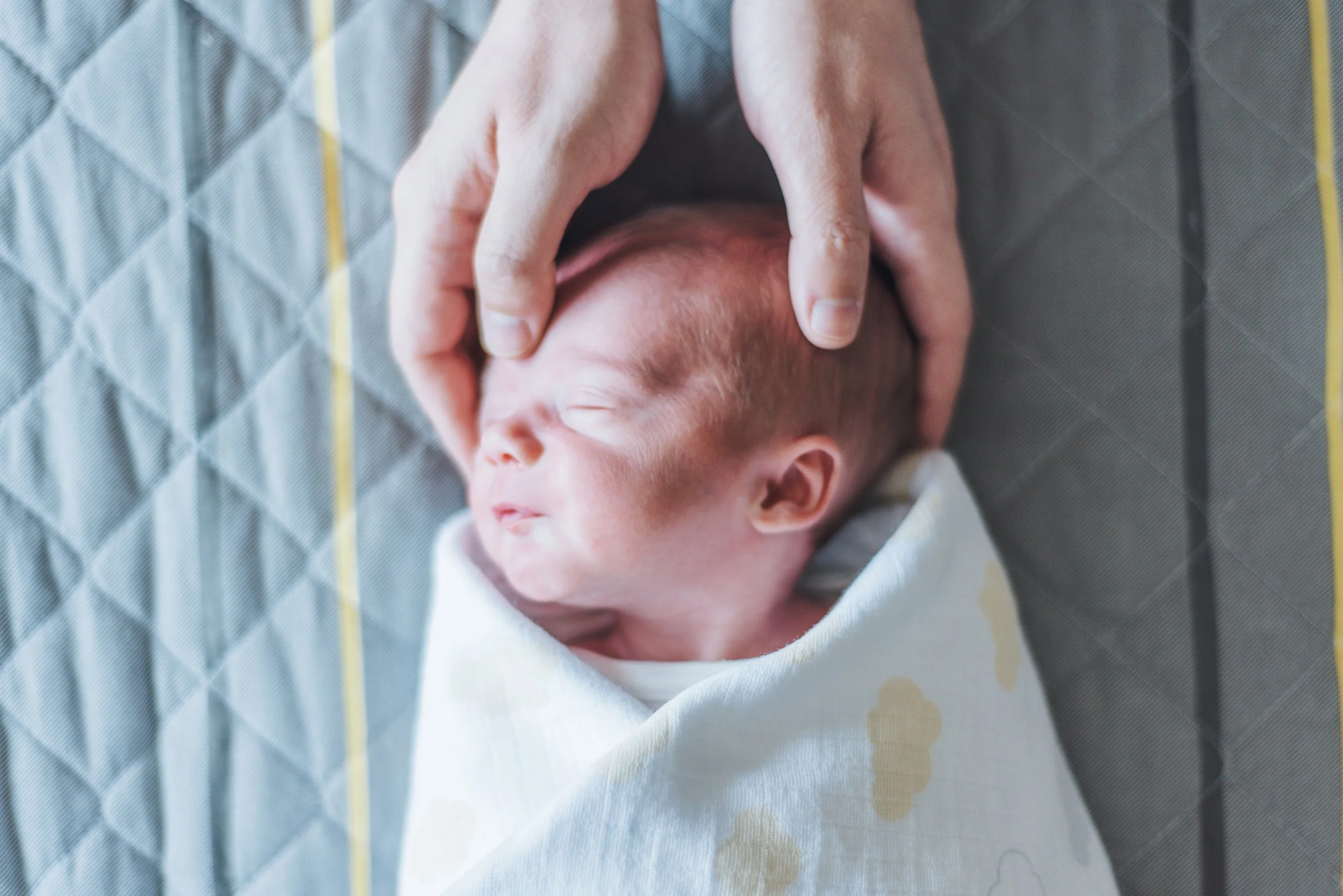 Close-up of a newborn baby wrapped in a white blanket with yellow cloud prints, sleeping peacefully with an adult's hands gently holding its head.