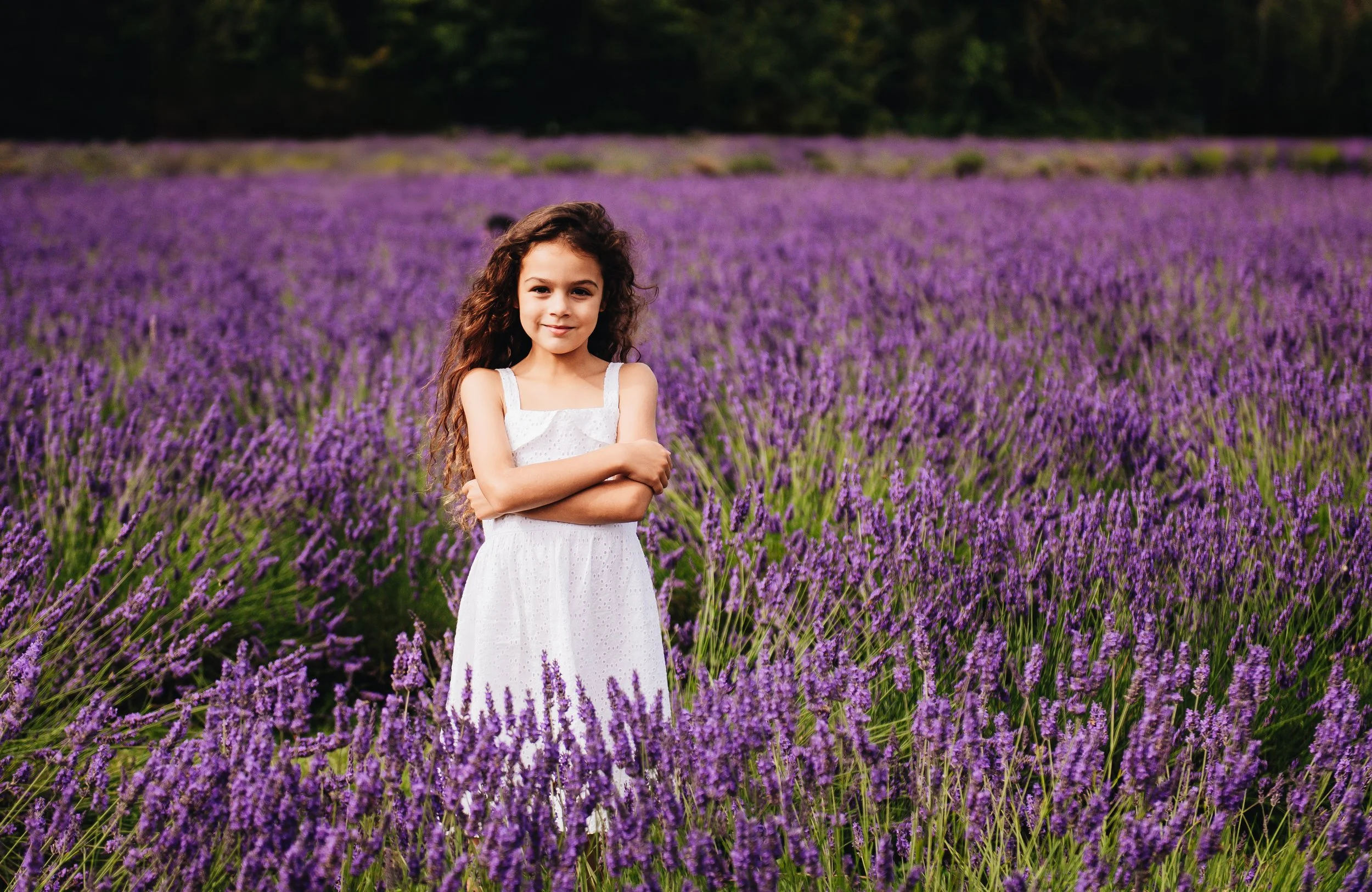 A young girl with long, curly brown hair in a white dress standing with arms crossed in a field of purple lavender flowers.