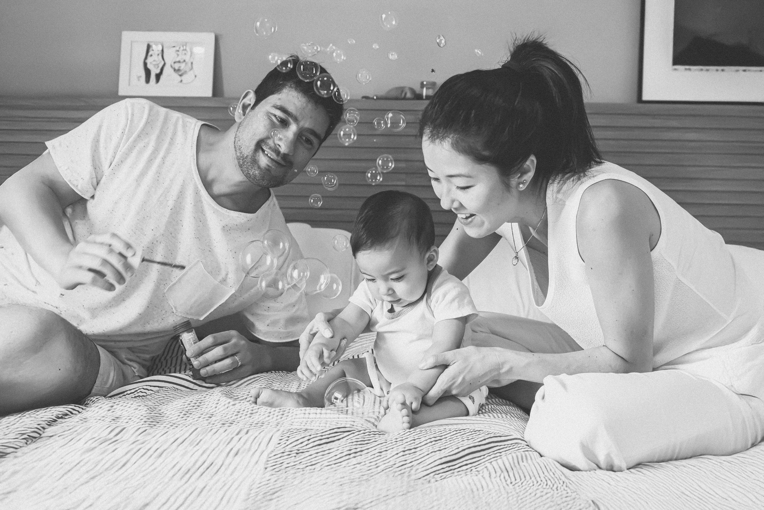 A family of three playing with soap bubbles on a bed, smiling and laughing, in a black and white photo.