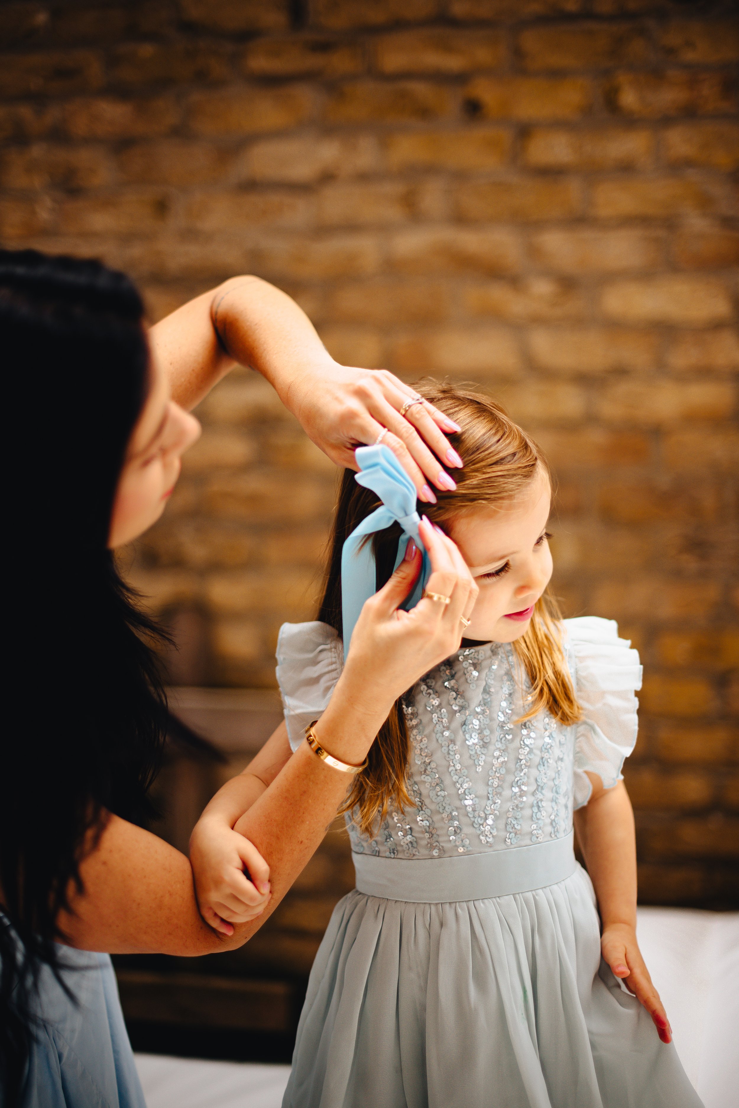 A woman styles a young girl's hair with a blue ribbon in a room with a brick wall.