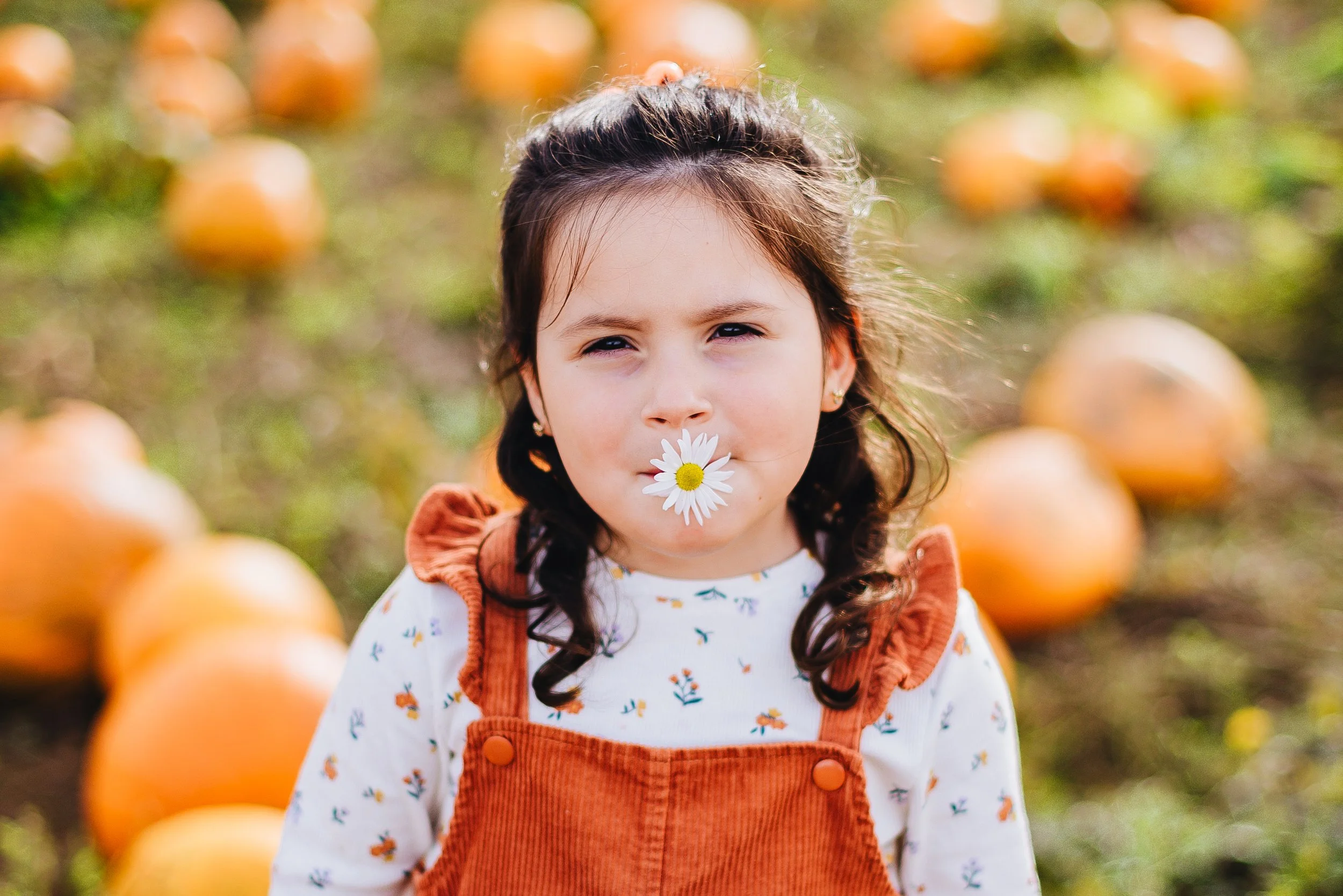 A young girl with dark hair, dressed in an orange overall and a white shirt with small floral prints, standing in a pumpkin patch with pumpkins in the background. She has a daisy flower in her mouth.