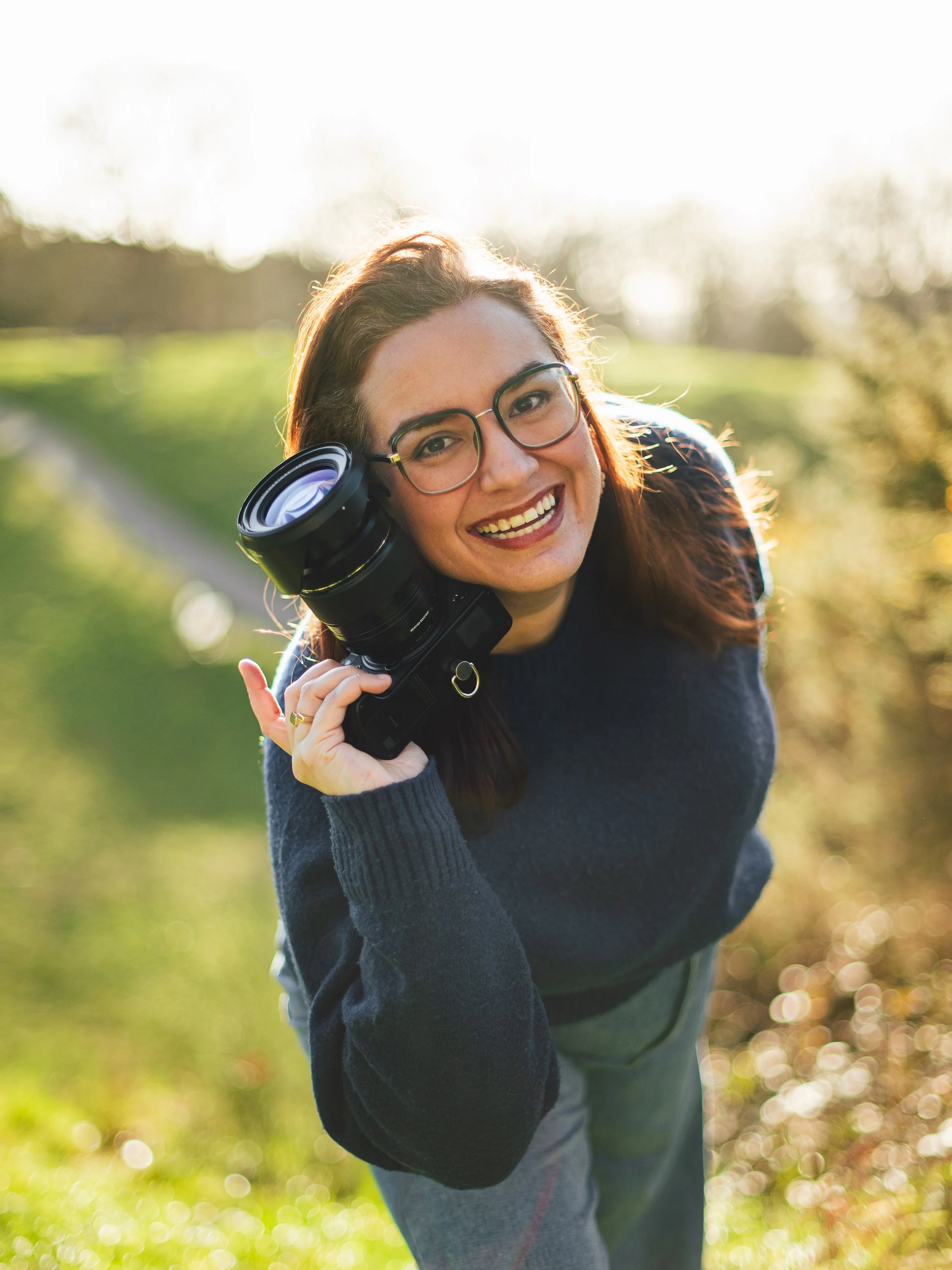 Woman smiling outdoors holding a professional camera with a large lens.