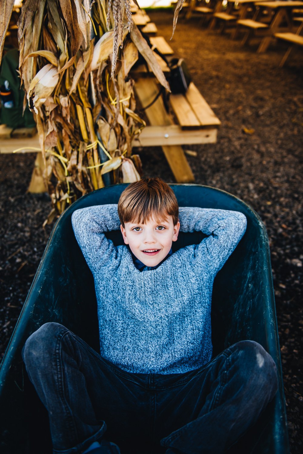 A young boy with brown hair, wearing a gray sweater and black jeans, lying back in a dark green wheelbarrow with his hands behind his head, outdoors on soil with a large bundle of dried cornstalks and a bench in the background.