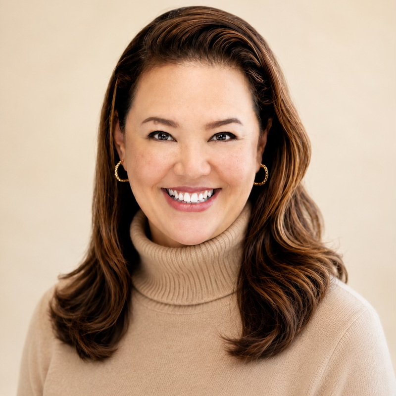 A woman with shoulder-length brown hair smiling and wearing a tan turtleneck sweater and gold hoop earrings.