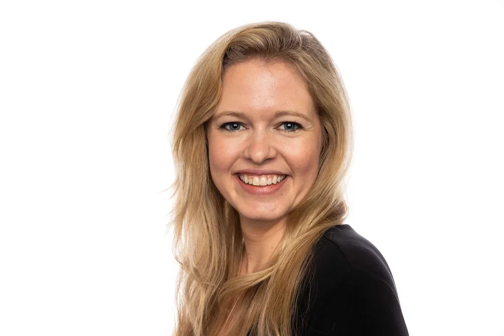 Portrait of a smiling young woman with long blonde hair wearing a black top against a white background.