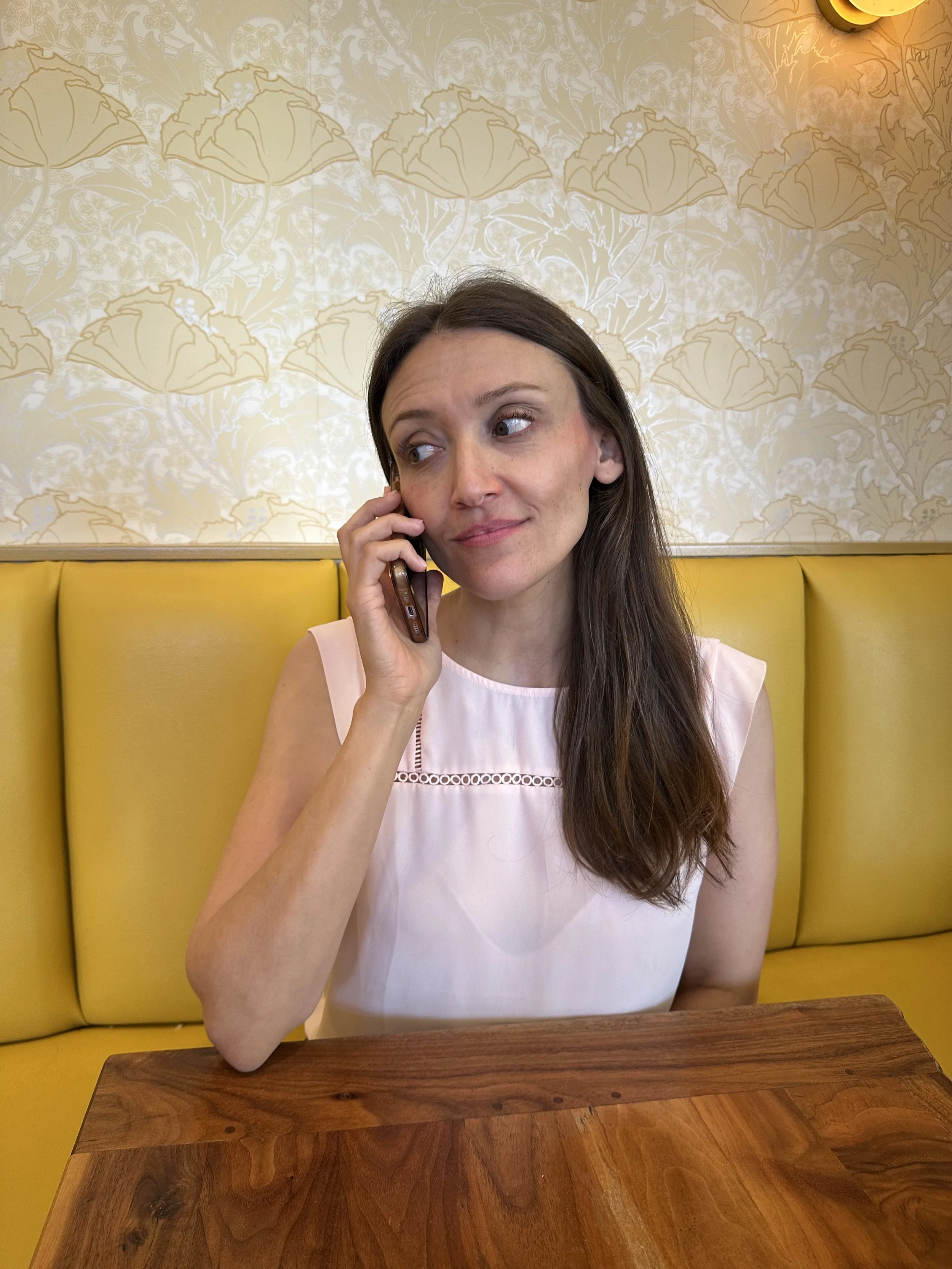 A woman with long brown hair, wearing a light pink sleeveless top, sitting at a wooden table, talking on a cell phone with a yellow cushioned booth and patterned wallpaper in the background.