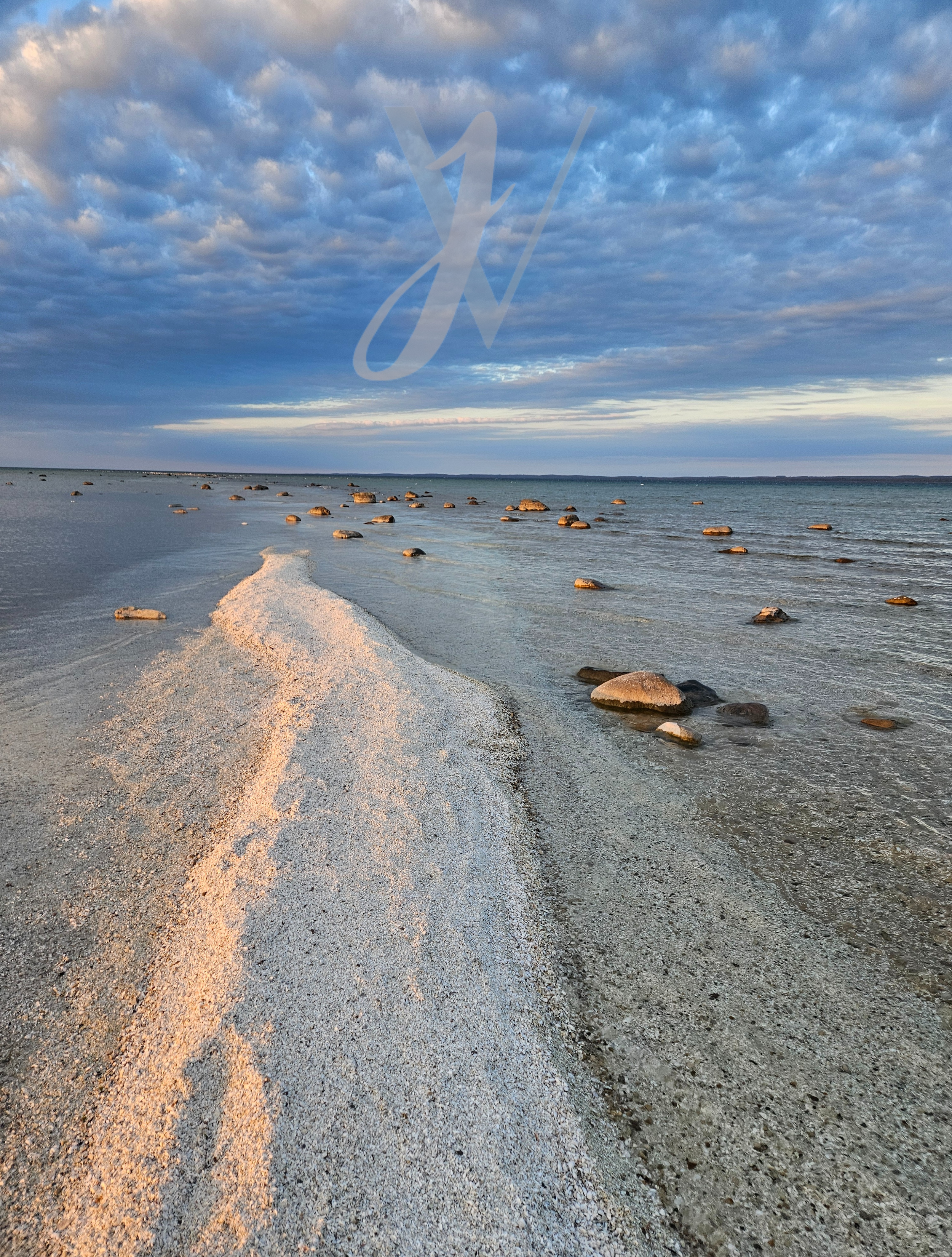 A beach with a narrow strip of sand and rocks extending into the water under a cloudy sky at sunset or sunrise.