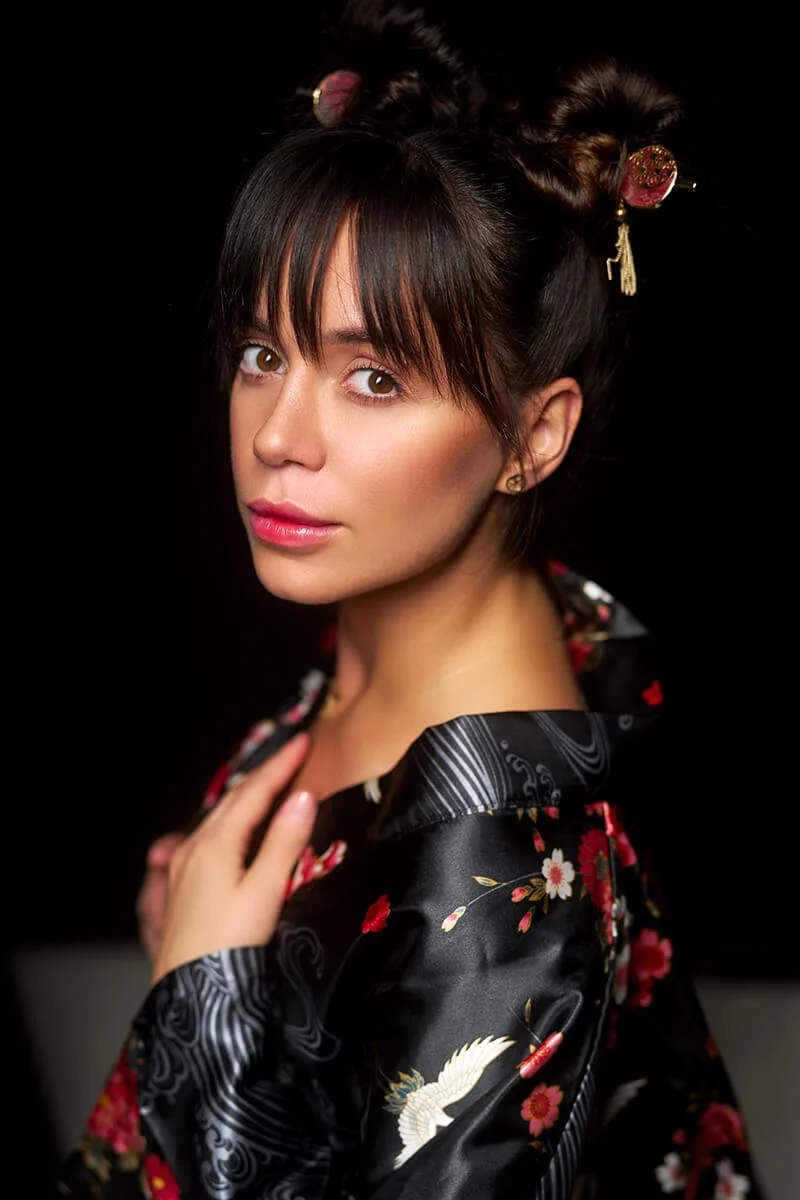 A woman with dark hair styled in traditional Japanese hair accessories, wearing a black kimono with a floral and crane pattern, looking at the camera against a black background.