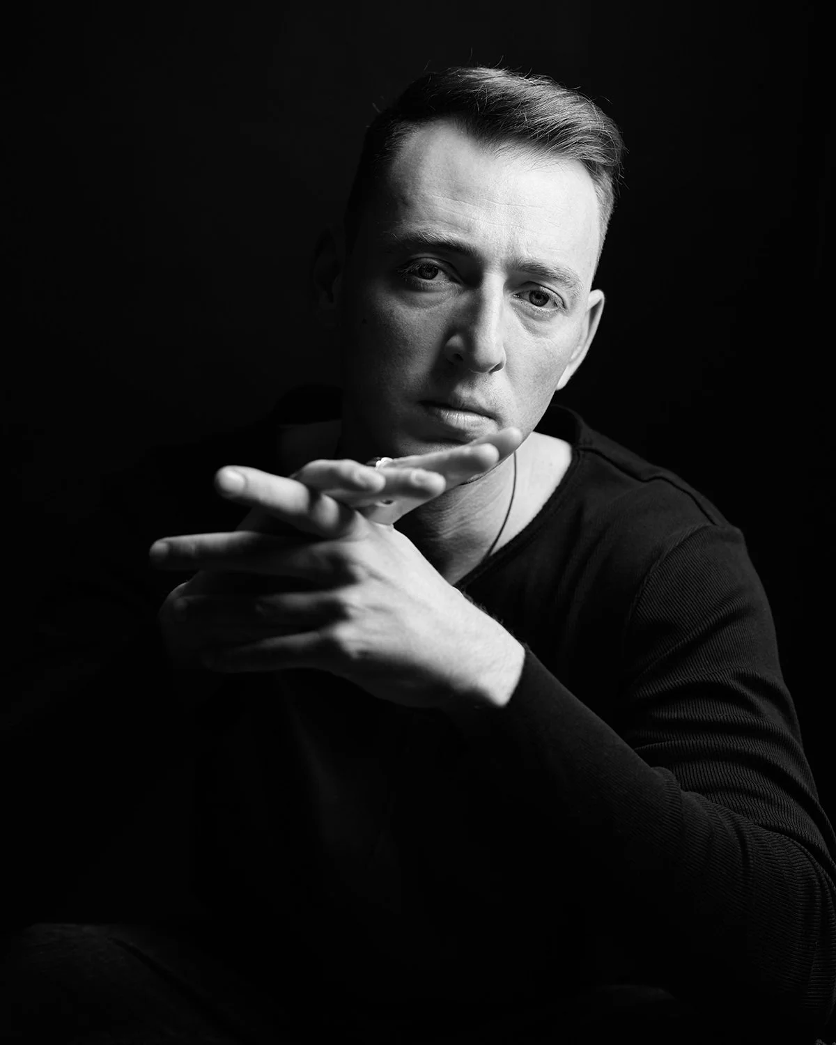 Black and white portrait of a young man with short hair, looking directly at the camera with a serious expression, resting his chin on his hand in front of a dark background.