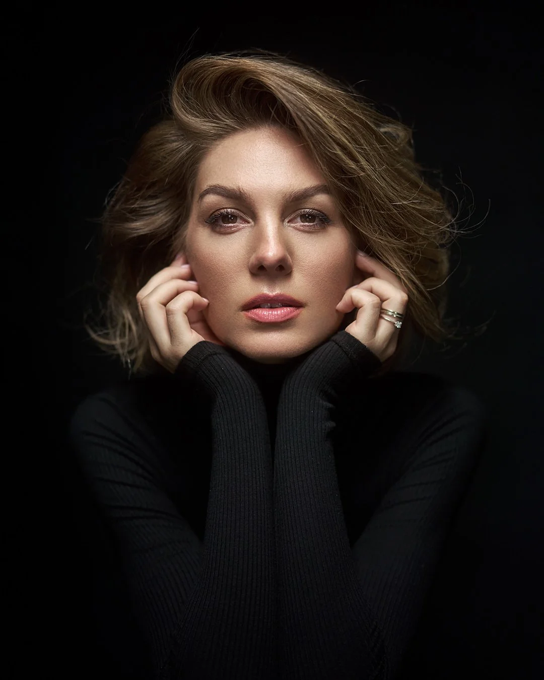 A portrait of a woman with wavy, shoulder-length brown hair, wearing a black turtleneck sweater, looking directly at the camera with her hands gently touching her face against a dark background.