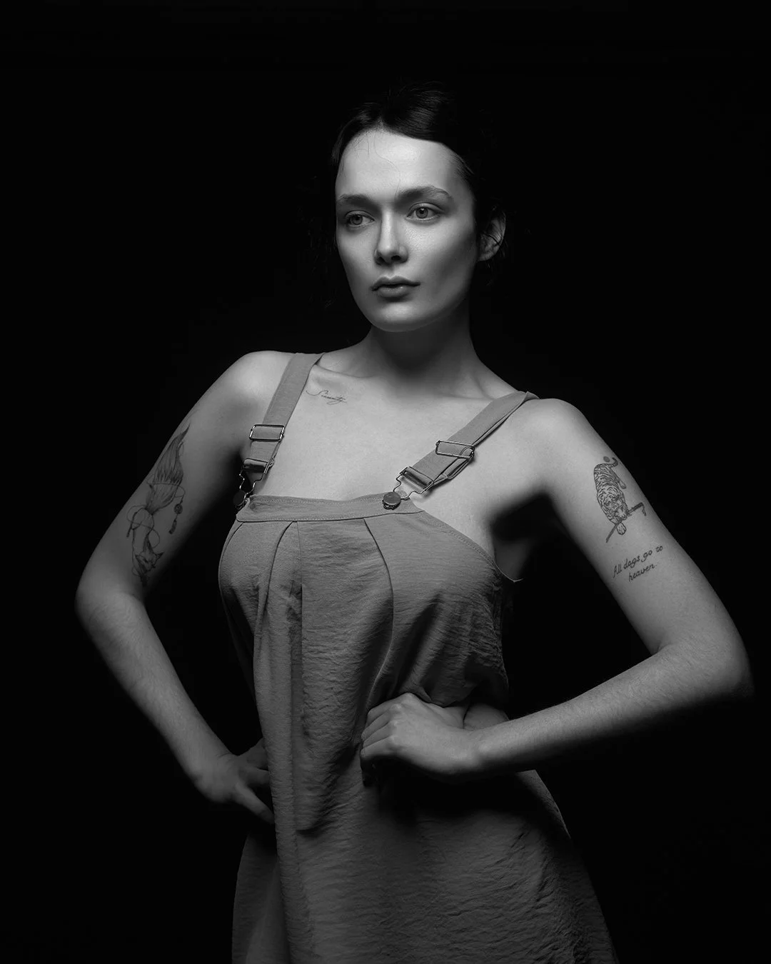 Femeie cu tatuaje pe brațe pozând cu mâinile în șolduri într-un studio foto, purtând o rochie cu bretele, iluminare dramatică pe fundal negru.