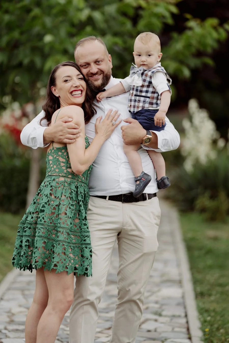 A smiling family of three standing on a stone path in a park, with green trees in the background. The woman is wearing a green lace dress, the man is wearing a white shirt, and the young boy is in a plaid vest and shorts, sitting on the man's arm.