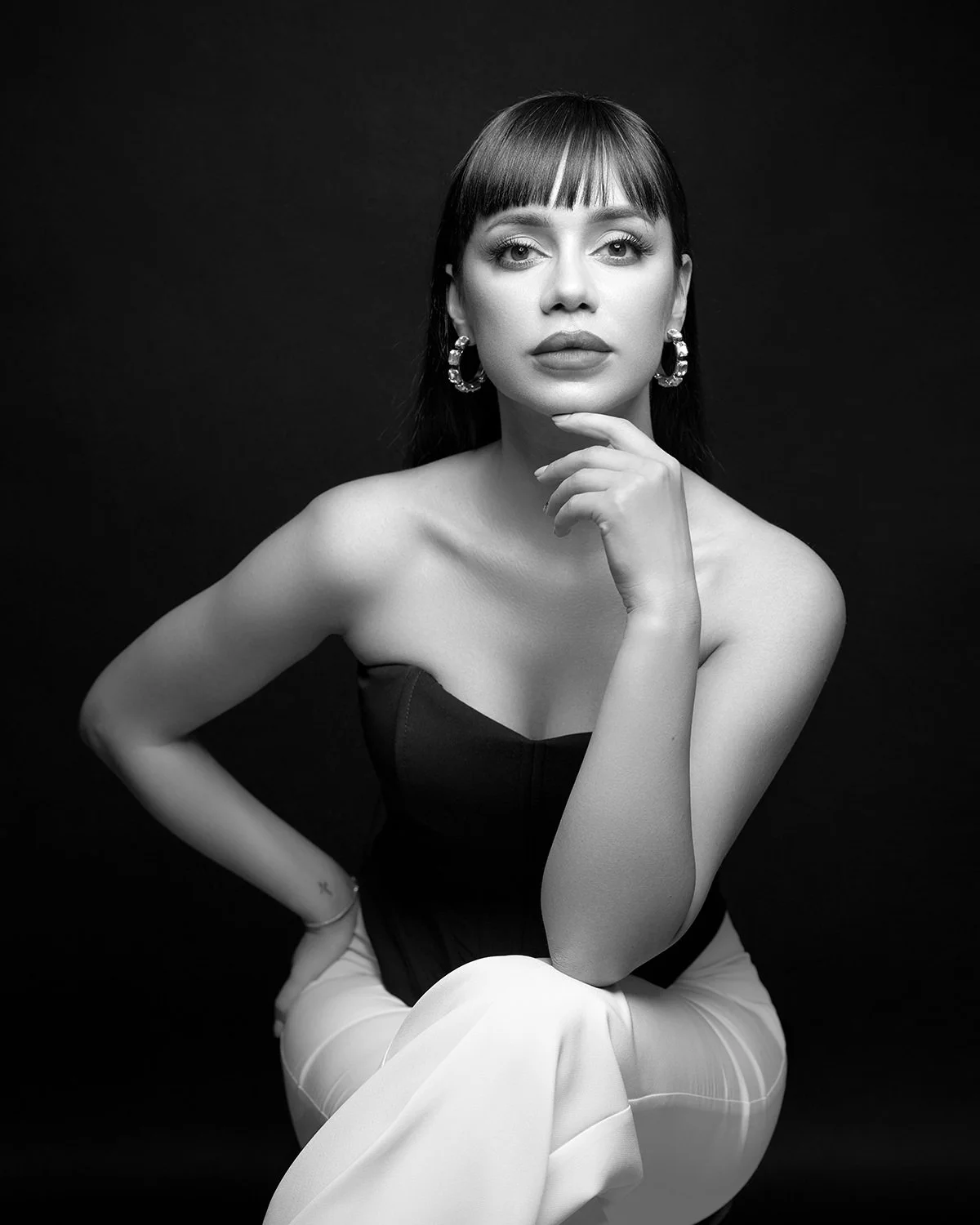 A black and white portrait of a woman with dark hair, wearing hoop earrings, a strapless top, and light-colored pants, posing with one hand under her chin and the other behind her back against a dark background.