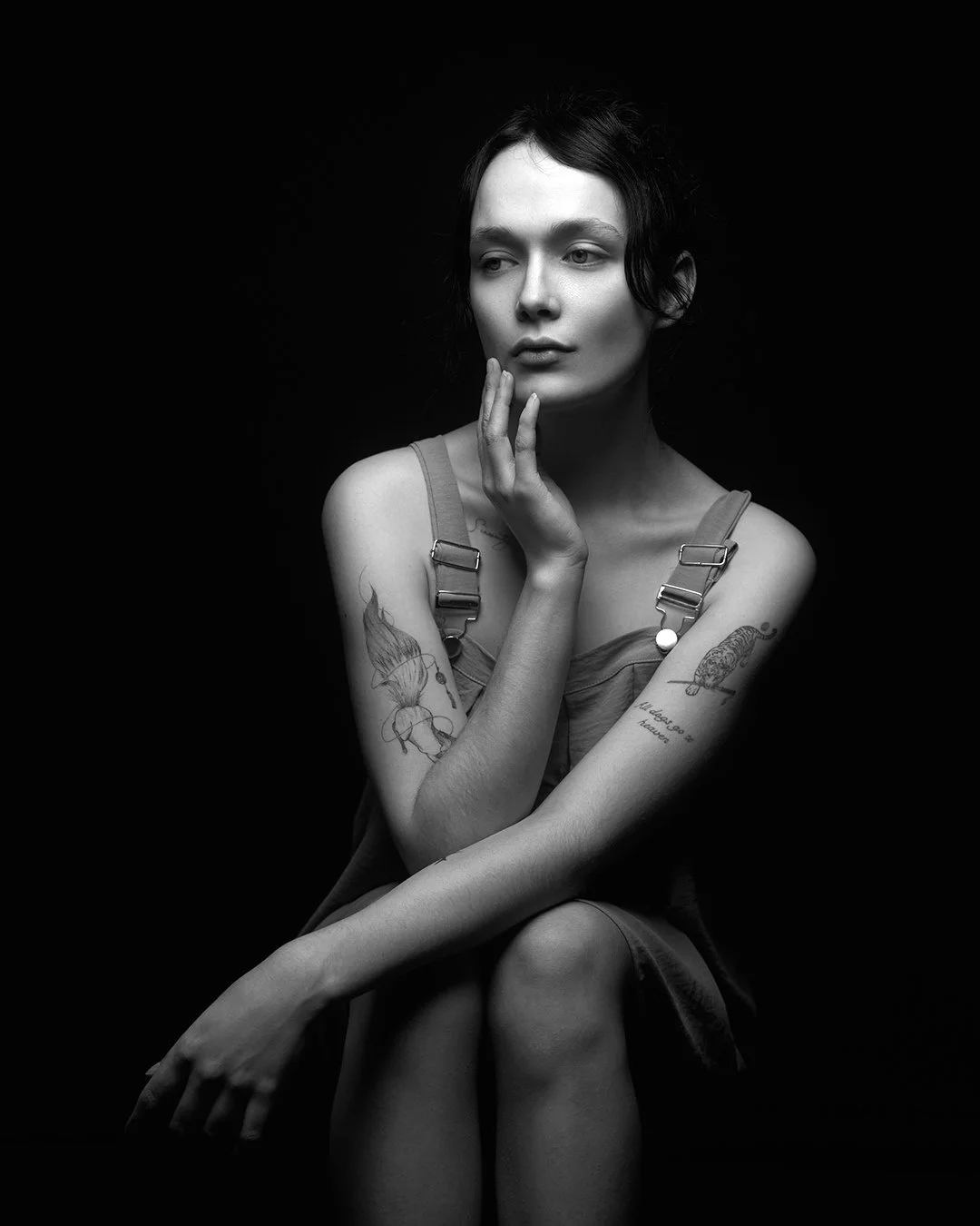 Black and white portrait of a woman with short dark hair, wearing a sleeveless top, sitting with her hand touching her face against a dark background.
