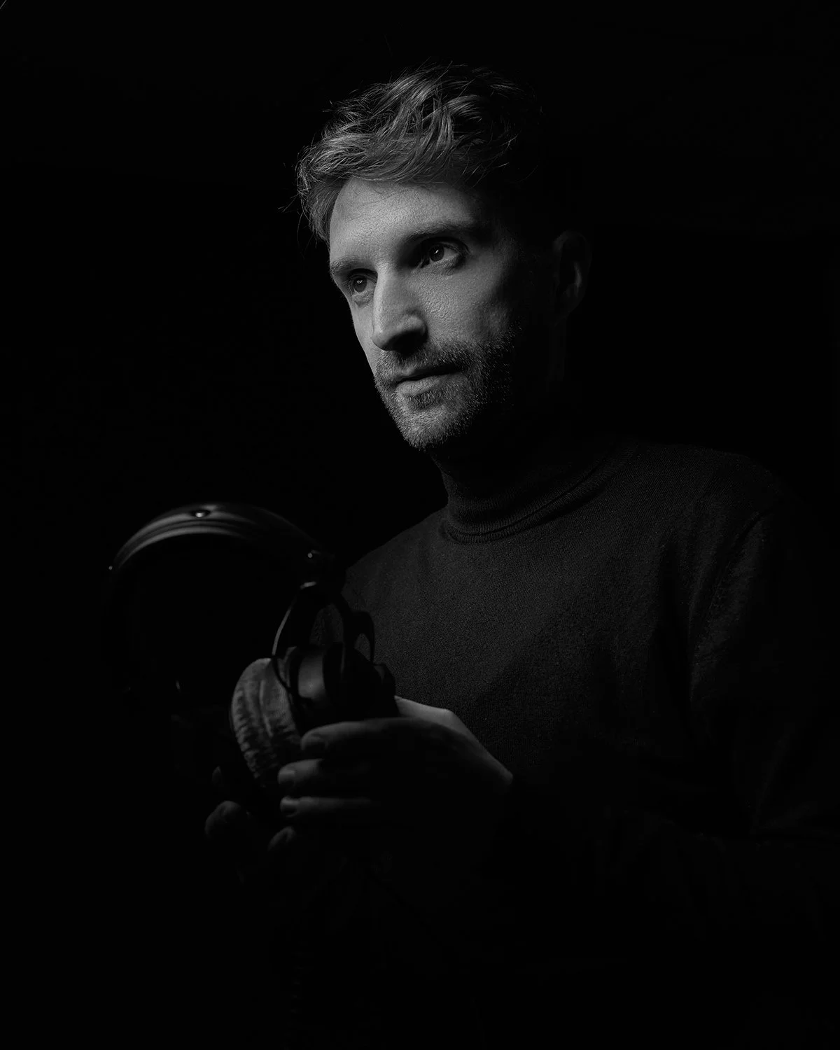Black and white portrait of a man with styled hair, holding a pair of headphones, looking to the side with a serious expression, dark background.