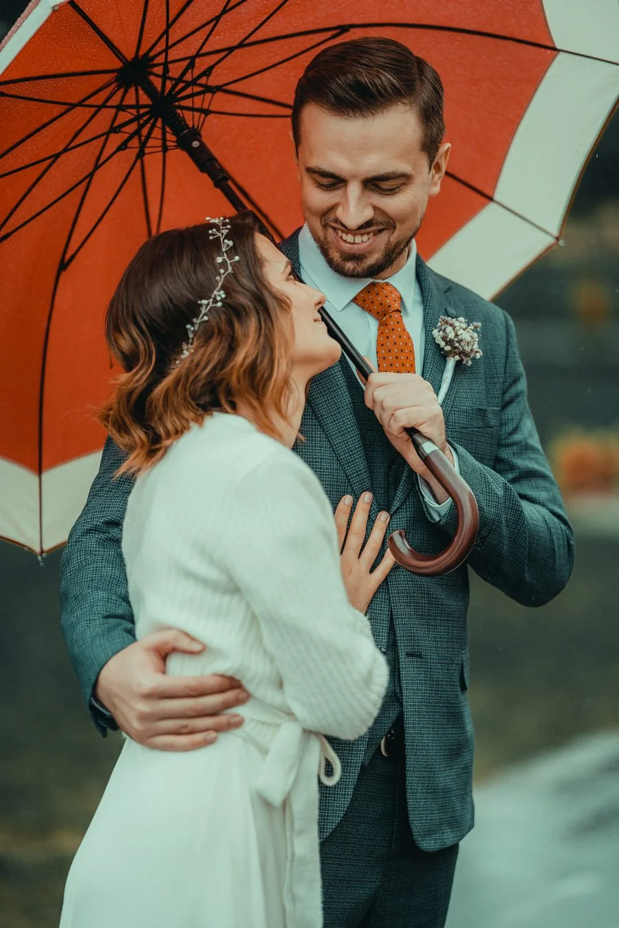 A couple at an outdoor wedding sharing a romantic moment under a red umbrella, with the groom smiling and holding the bride close.