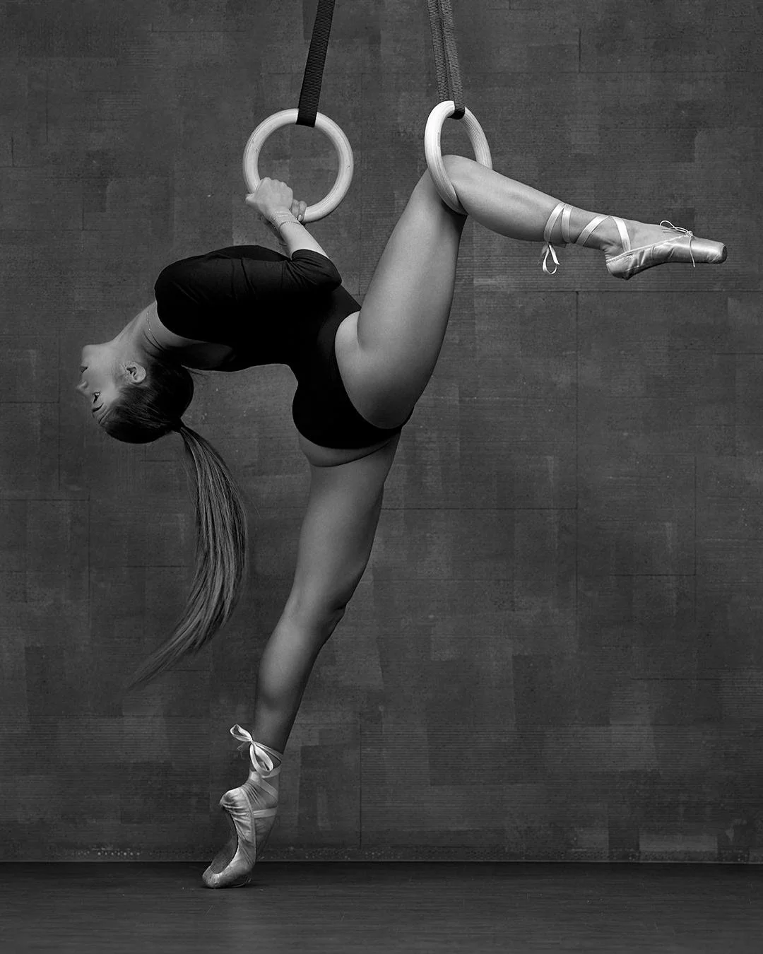 Ballet dancer performing an aerial pose on gymnastics rings, with one leg extended and pointe shoes, in a studio with wooden floor and wall.