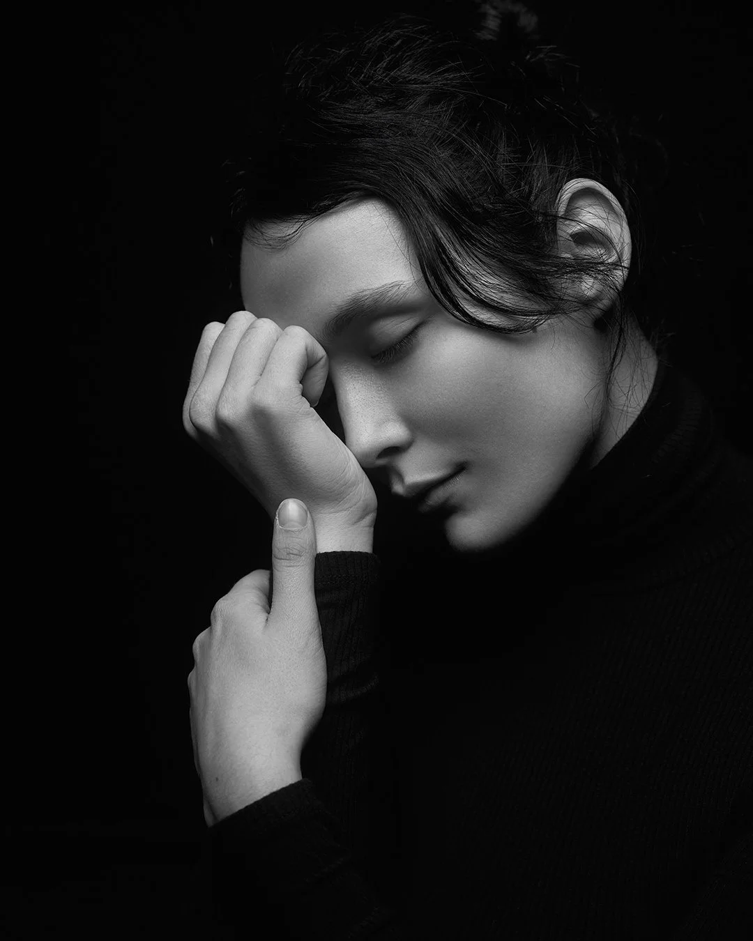 Black and white portrait of a woman with closed eyes, resting her forehead on her hand, wearing a black turtleneck.