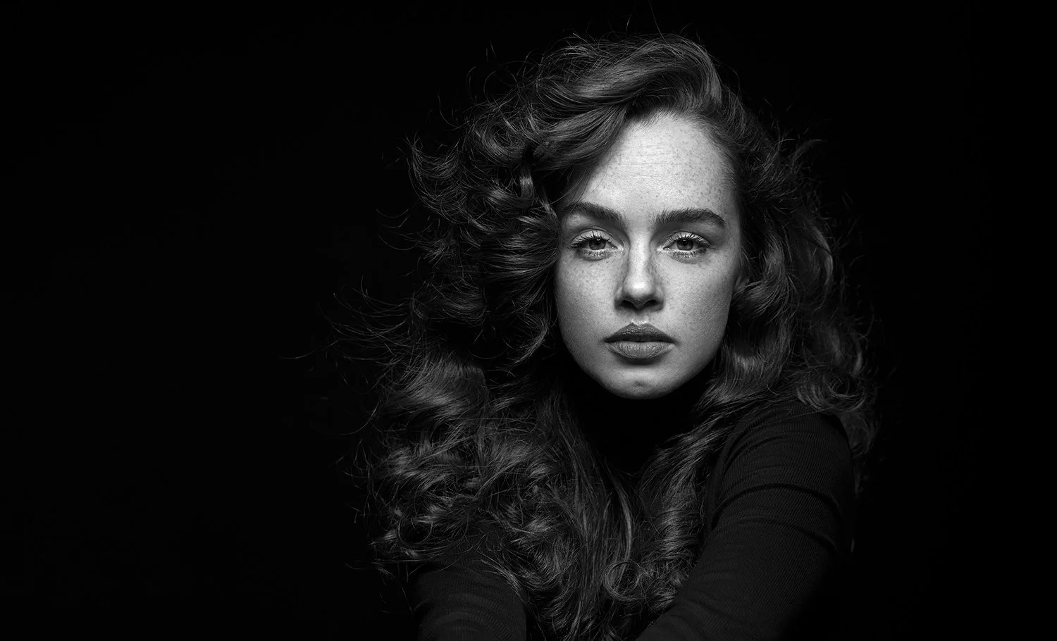 Portret close-up alb-negru al unei femei cu pistrui si par ondulat, privire intensa, realizat in studio cu iluminare dramatica pe fundal negru.