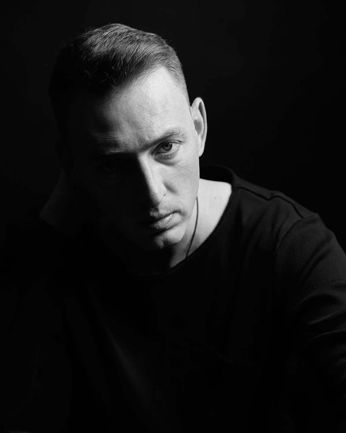 A black and white portrait of a man with short hair, looking directly at the camera with a serious expression, dark background, wearing a dark shirt.