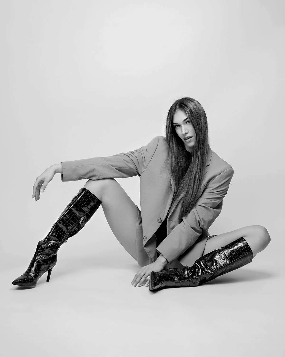 Black and white photo of a woman sitting on the floor, wearing a blazer and high-heeled boots, with long hair and an intense expression.