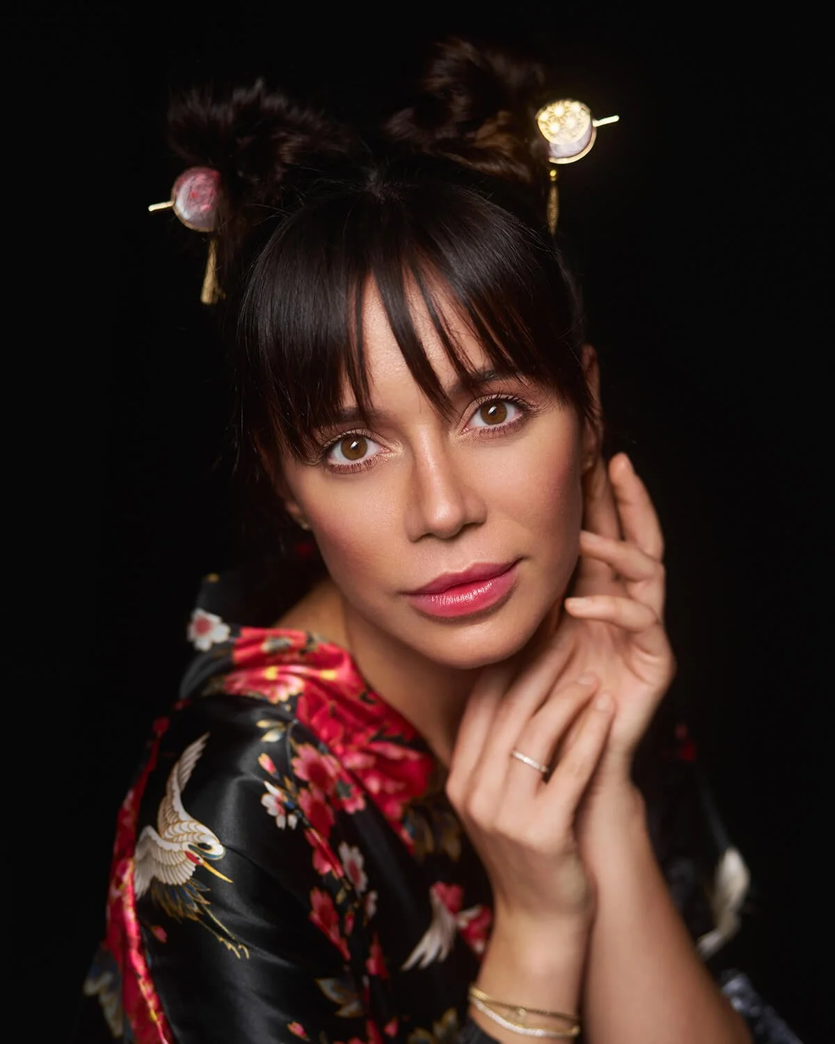 Portrait of a young woman with brown hair styled in buns with hair sticks, wearing a black floral kimono, and posing against a black background.