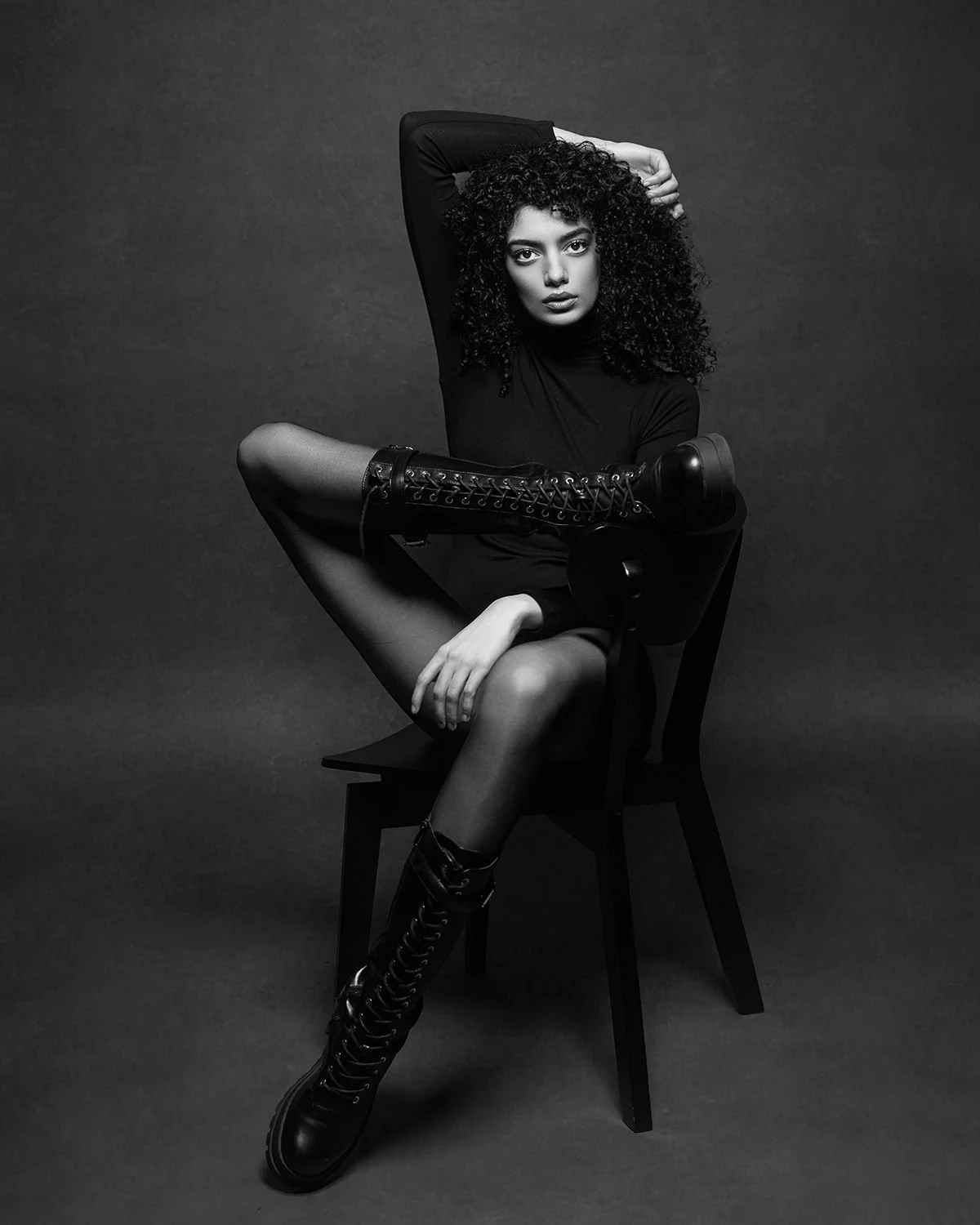 Black and white photo of a woman with curly hair sitting on a chair with one leg on the seat. She is wearing a long-sleeved black top, sheer tights, and tall, lace-up boots. She has one arm over her head and is looking directly at the camera.