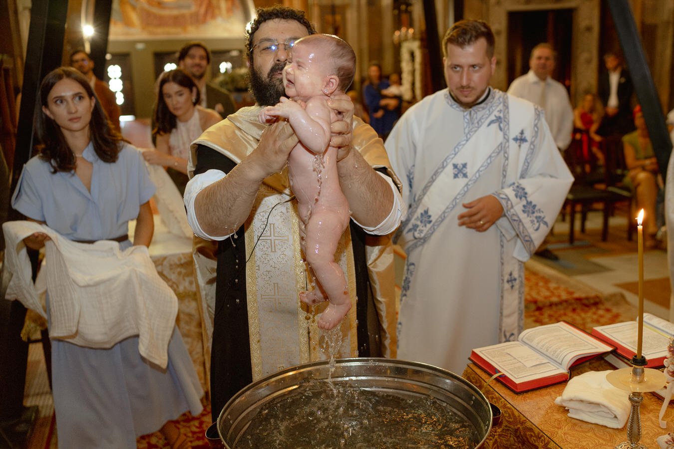 A priest holding a baby during a baptism ceremony in a church, with people watching in the background.