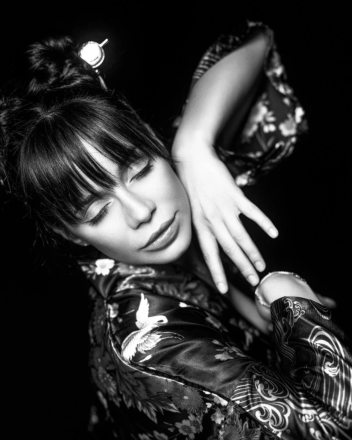 A woman with dark hair and bangs, wearing traditional clothing with floral and crane patterns, poses with her eyes closed and her hand resting on her neck, against a black background.