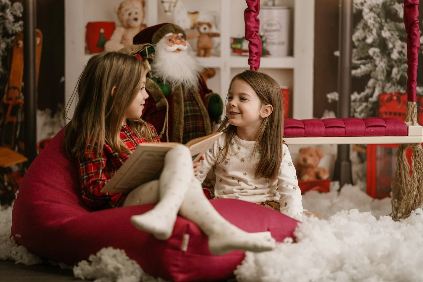 Two young girls sitting on the floor, one reading a book, in a Christmas decorated room with Santa Claus figure and teddy bears in the background.