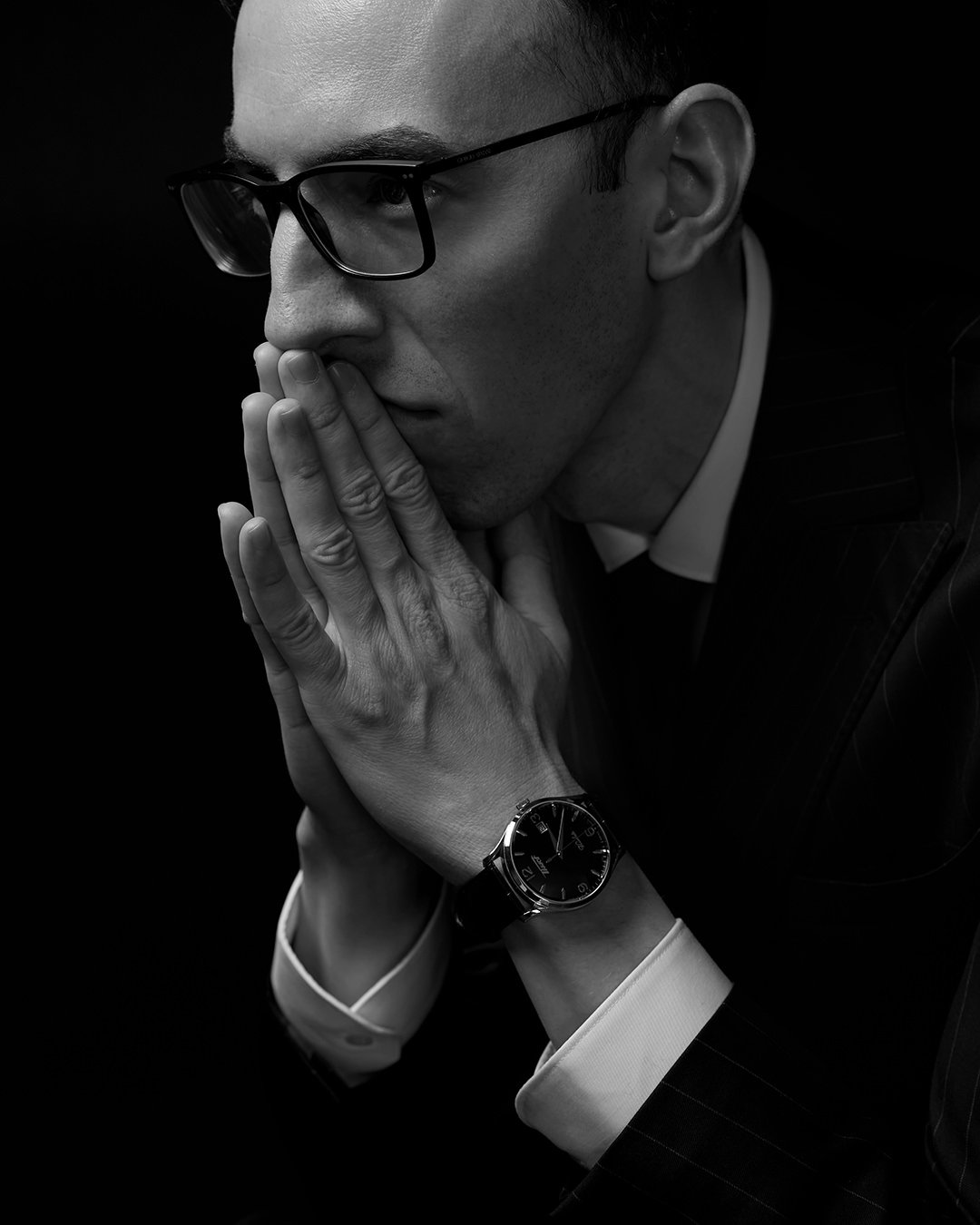 Black and white portrait of a man with glasses, bald head, wearing a watch and business attire, with hands clasped near his face in a thoughtful pose.
