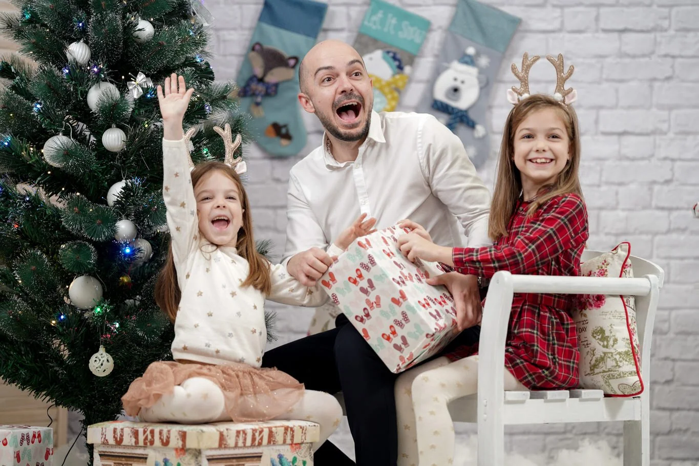 A man with two young girls celebrating Christmas indoors, exchanging gifts near a decorated Christmas tree with ornaments and lights, with stockings hanging on the wall in the background.
